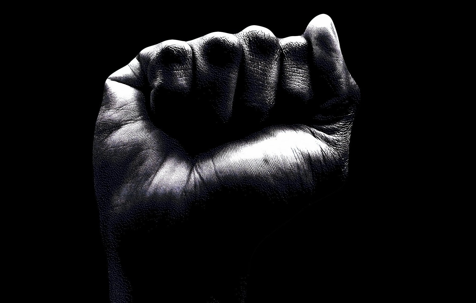 L'image représente un poing fermé, vu de près sur un fond noir. La main est d'une teinte sombre, avec des détails visibles sur la peau, comme les lignes des empreintes digitales et les plis. Le poing, symbolisant souvent la force ou la résistance, est levé de manière à exprimer la détermination. Les contours de la main sont nets, mettant en avant la texture et la forme. L'arrière-plan noir accentue la présence du poing, créant un contraste fort et saisissant.