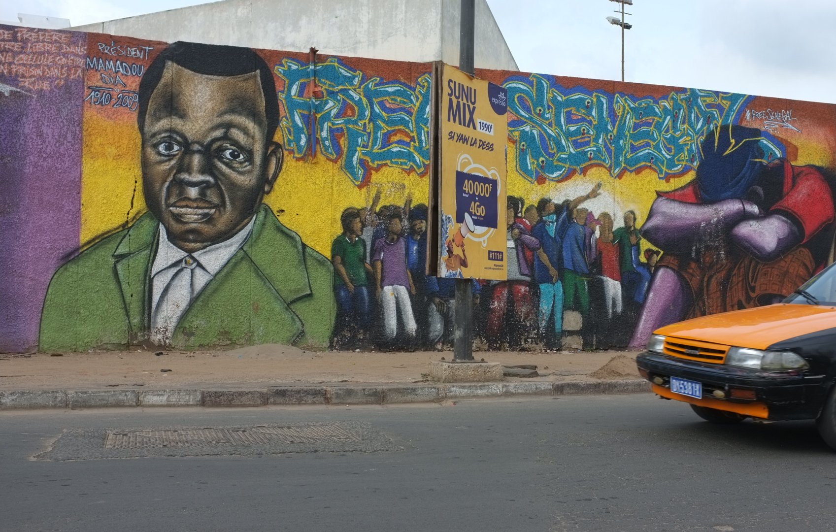 L'image montre un mur de graffitis vibrant et coloré. Au centre, il y a un portrait d'une personne au visage sérieux, avec des traits marqués, habillée d'un costume vert. Autour de ce portrait, différentes figures sont représentées, suggérant une scène animée de personnes rassemblées, peut-être en train de discuter ou de célébrer. À côté, on peut voir une affiche collée sur le mur avec des éléments graphiques. En bas de l'image, un taxi à la carrosserie orange et noir passe sur la route, ajoutant une touche de dynamisme à la scène urbaine. L'ensemble évoque une ambiance de vie communautaire et de culture urbaine.