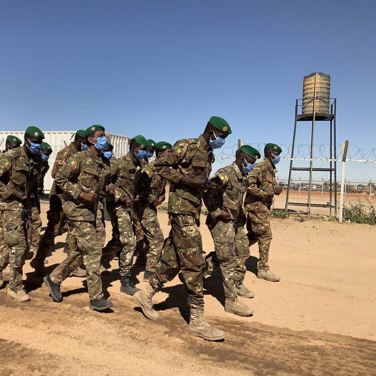 Dans cette image, un groupe de soldats est en train de marcher en formation sur un chemin poussiéreux. Ils portent des uniformes militaires verts camouflés et des casquettes vertes. La majorité d'entre eux portent également des masques bleus, ce qui suggère une mesure de protection sanitaire. En arrière-plan, on peut distinguer des conteneurs et une tour d'eau. Le ciel est dégagé, avec une couleur bleue intense, créant un contraste avec la terre orange et les silhouettes des soldats. L'atmosphère évoque un moment de discipline et d'organisation dans un environnement militaire.