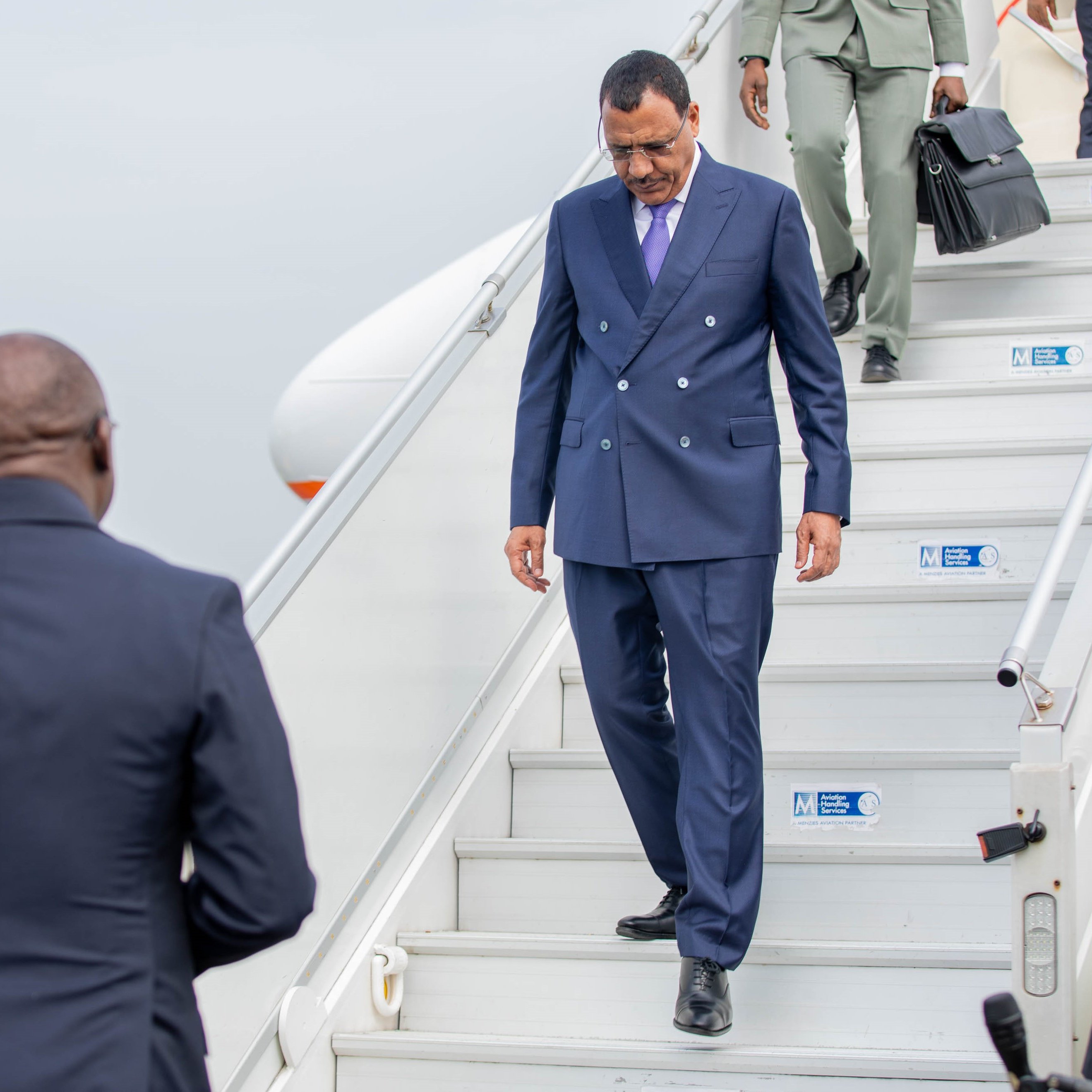 L'image montre un homme qui descend des marches d'un avion. Il porte un costume bleu foncé et des lunettes de soleil. Son expression semble concentrée tandis qu'il s'avance avec assurance. À côté de lui, un autre homme en costume noir observe attentivement. En arrière-plan, on aperçoit des membres du personnel de l'aéroport, ainsi que l'avion stationné. L'atmosphère peut sembler formelle et dynamique, suggérant un moment important, vraisemblablement une arrivée ou une sortie officielle.