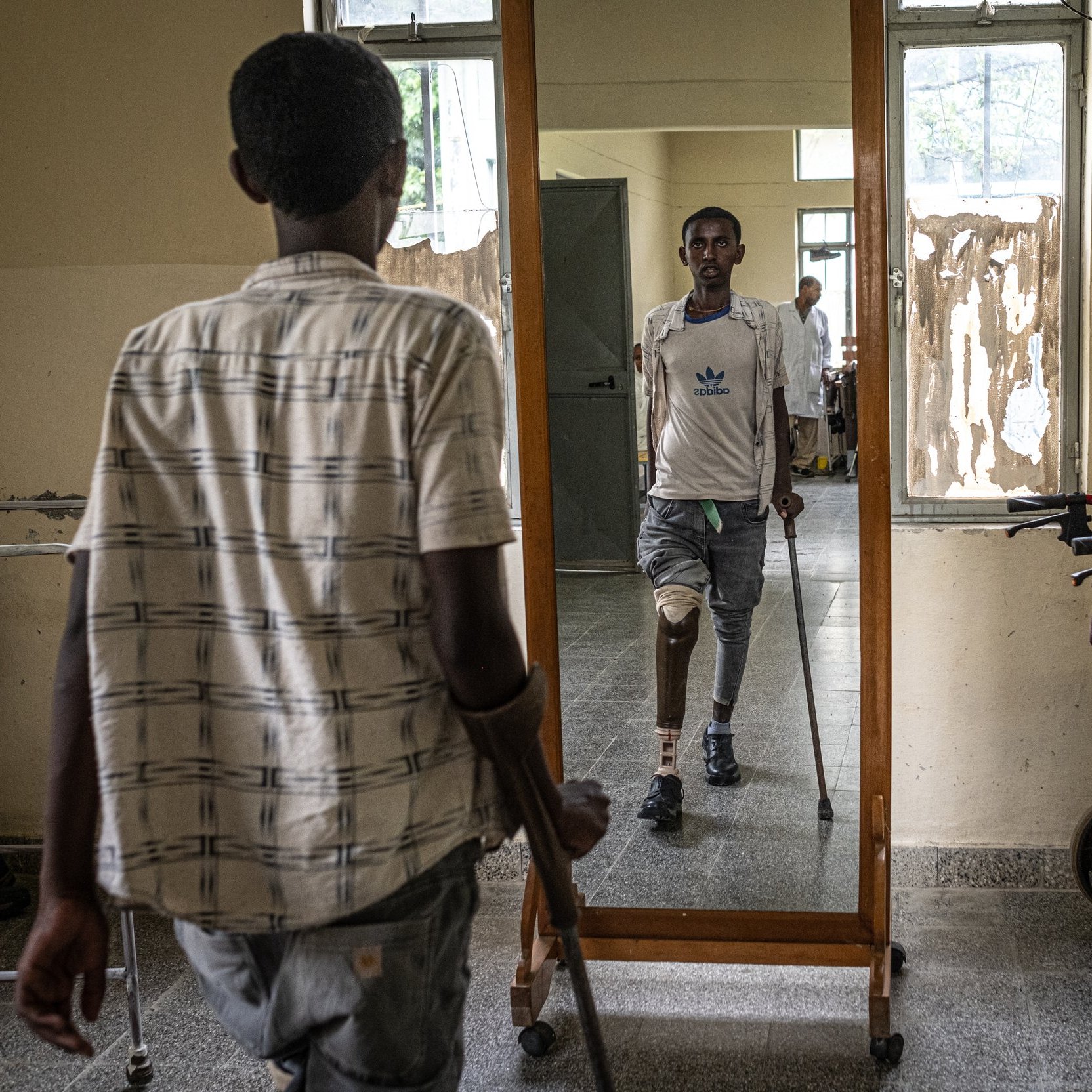 L'image montre une scène dans un lieu qui semble être un centre de réhabilitation ou un hôpital. Au premier plan, un jeune homme se tient à l'aide de béquilles, son regard fixé sur son reflet dans un grand miroir devant lui. Il porte une chemise à rayures et des pantalons clairs. Dans le miroir, on peut voir un autre homme, qui a une prothèse de jambe et se tient debout, semblant plus confiant. Sur le côté, une chaise roulante est disposée, suggérant un environnement d'assistance. Les murs autour d'eux sont simples et montrent des signes d'usure. L'atmosphère évoque des efforts de réhabilitation et de courage.