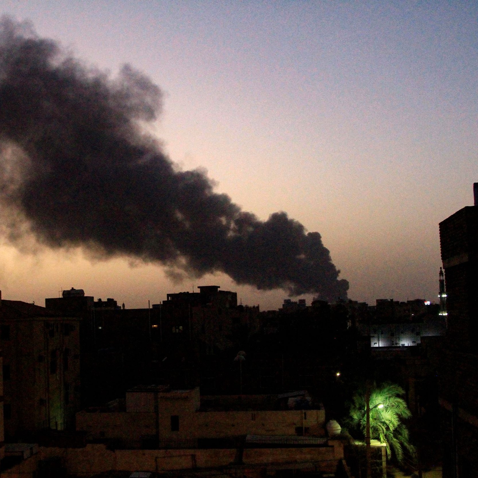 L'image montre un paysage urbain au crépuscule, où le ciel est teinté de nuances de bleu et d'orange. En arrière-plan, une épaisse colonne de fumée noire s'élève, suggérant une source d'incendie. Les silhouettes de bâtiments se dessinent en avant-plan, avec des structures variées et quelques lumières allumées. À droite, on aperçoit un palmier, ajoutant une touche de verdure à ce paysage sombre et chargé d'une ambiance d'urgence.
