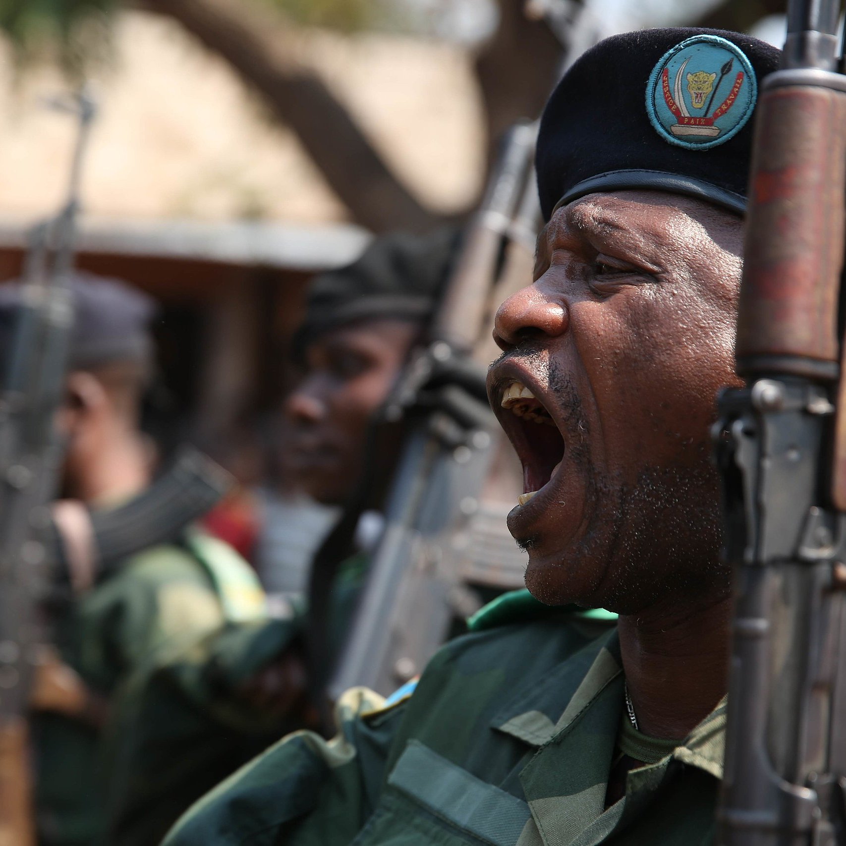 L'image montre un groupe de militaires en uniforme, en position de parade. L'un d'eux, au premier plan, est en train de crier ou de donner un commandement, affichant une expression de détermination. Ils portent des bérets sombres et tiennent des fusils en position verticale. En arrière-plan, on peut apercevoir d'autres soldats ainsi qu'un groupe de personnes qui semblent regarder la scène. L'environnement est extérieur, probablement lors d'un événement officiel ou d'une cérémonie. L'atmosphère est solennelle et dynamique, évoquant un sentiment de force et de discipline.