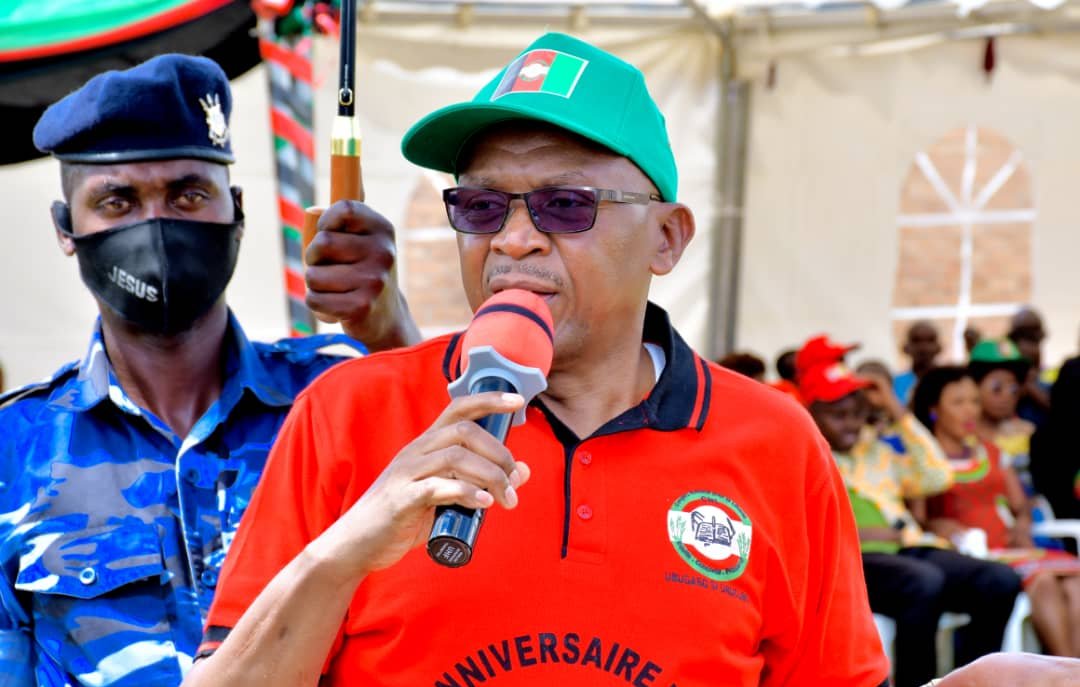 L'image montre un homme qui prend la parole devant un public. Il porte un t-shirt rouge avec le texte "ANNIVERSAIRE DU PARTI" et une casquette verte. Il tient un micro et semble s'adresser à l'assemblée avec passion. À l'arrière-plan, on peut apercevoir des gens assis, certains portant des vêtements colorés. Un agent de sécurité en uniforme bleu est visible à côté de lui, ajoutant un élément de protection ou d'autorité à l'événement. L'atmosphère semble festive et engagée.