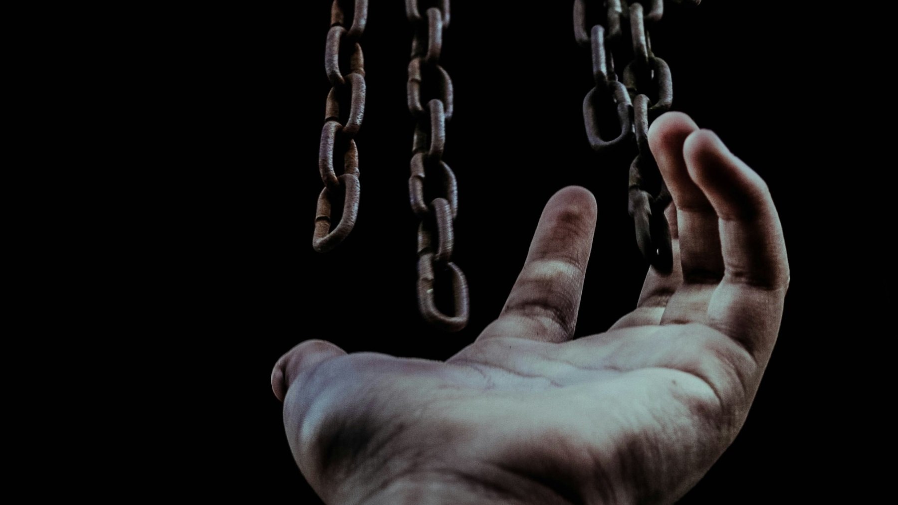 L'image montre une main tendue vers le haut, en direction de chaînes suspendues qui descendent. La main, légèrement ouverte, semble chercher à atteindre les chaînes, qui sont bien visibles sur un fond sombre. Les chaînes sont métalliques, avec une texture rustique, et elles évoquent une sensation d'emprisonnement ou de lutte. L'éclairage met en valeur les détails des chaînes et de la main, créant une atmosphère à la fois troublante et poignante. Cette composition suggère un thème de quête ou de désir de liberté.