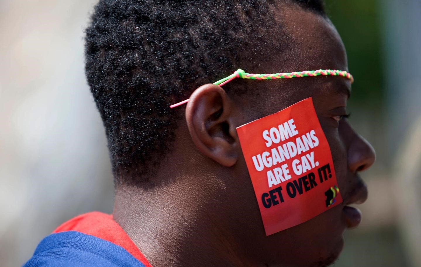 L'image représente un homme dont le visage est de profil. Il porte un petit autocollant sur sa joue qui affiche un message en lettres blanches sur fond rouge. Le texte dit : "SOME UGANDEANS ARE GAY. GET OVER IT!" (Certains Ougandais sont gays. Acceptez-le !). L'homme a des cheveux courts et un accessoire coloré autour de la tête. L'ambiance générale de l'image suggère un acte de protestation ou de revendication des droits.