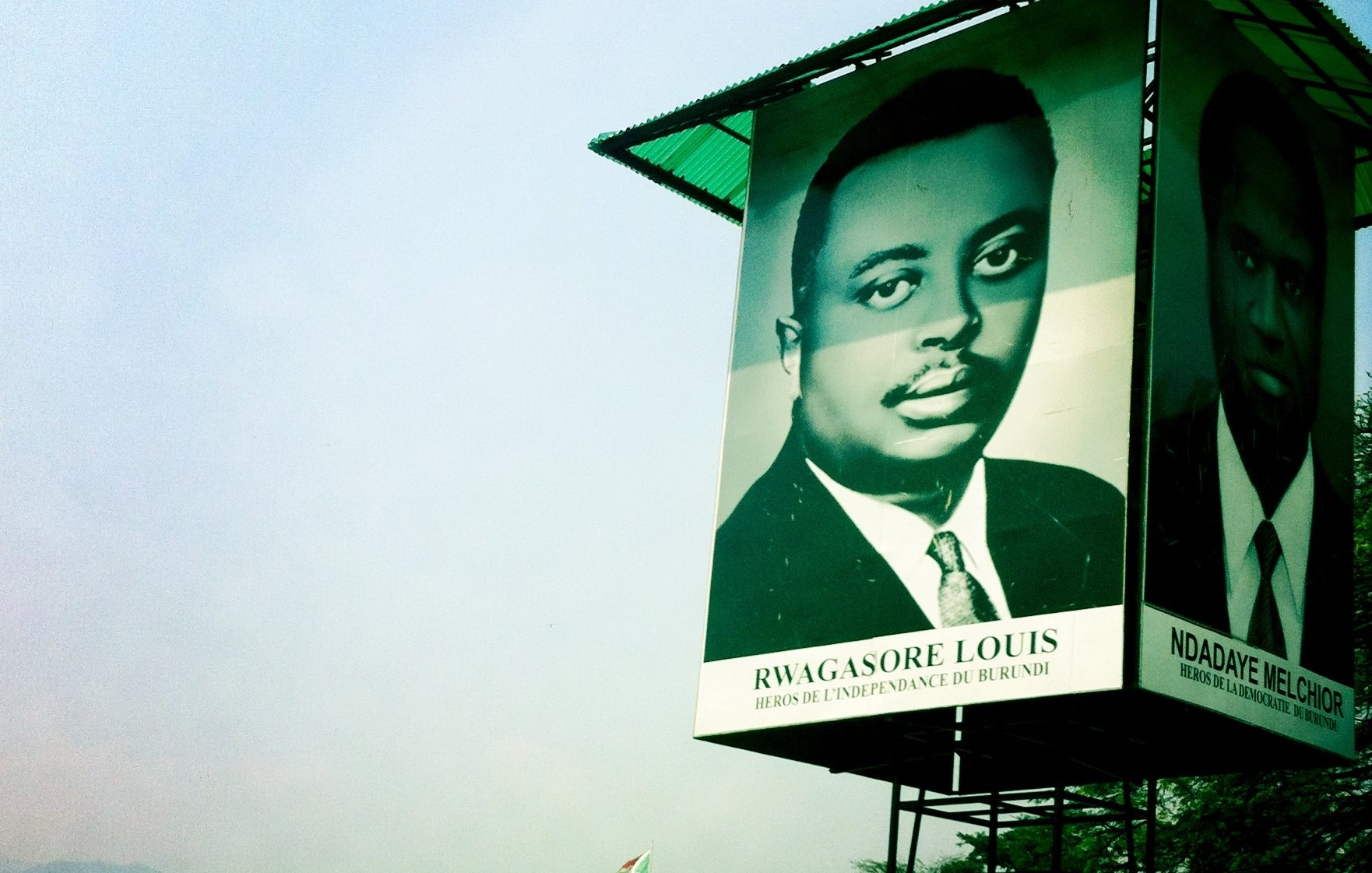 Cette image montre un grand panneau publicitaire ou une affiche située dans un environnement urbain. Sur le panneau, il y a un portrait en noir et blanc d'un homme, sous lequel est inscrit son nom : "RWAGASORE LOUIS", avec la mention "Héros de l'indépendance du Burundi". À côté de ce portrait, on aperçoit un autre visage (sans nom visible). Le fond de l'image présente un ciel légèrement brumeux et des bâtiments en arrière-plan, ce qui indique un paysage urbain. L'ambiance de l'image est à la fois historique et commémorative, mettant en avant des figures importantes pour le Burundi.