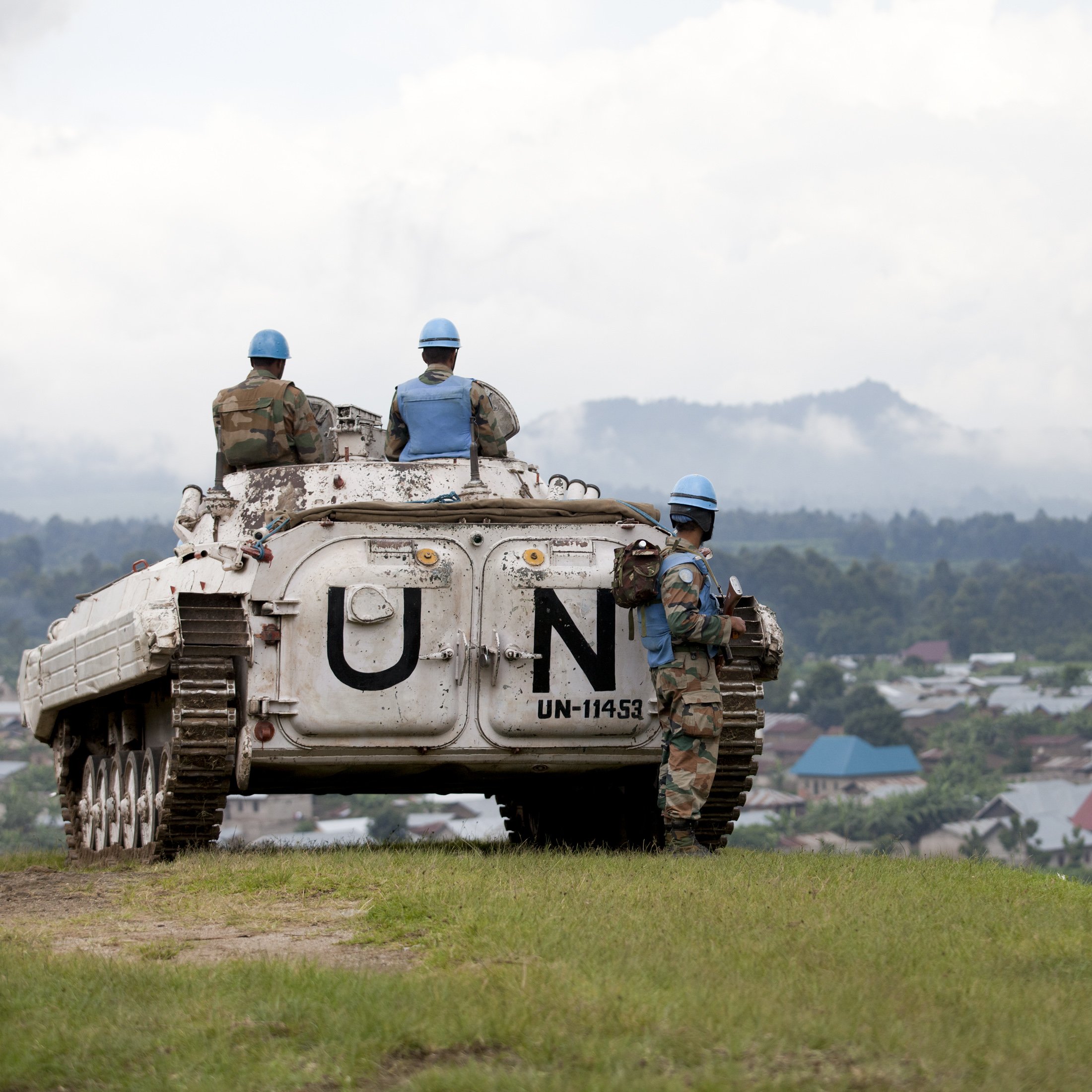 The image depicts a scene from a military operation involving United Nations peacekeepers. In the foreground, a military vehicle marked with "UN" is parked on a grassy area overlooking a landscape. Several soldiers in blue helmets, which signify their affiliation with the UN, are positioned around the vehicle. They appear to be surveying the area, which includes a view of a village with rooftops in the background. The atmosphere seems calm, with a cloudy sky adding to the setting. The soldiers' uniforms suggest they are part of a peacekeeping mission, emphasizing their role in maintaining stability in the region.