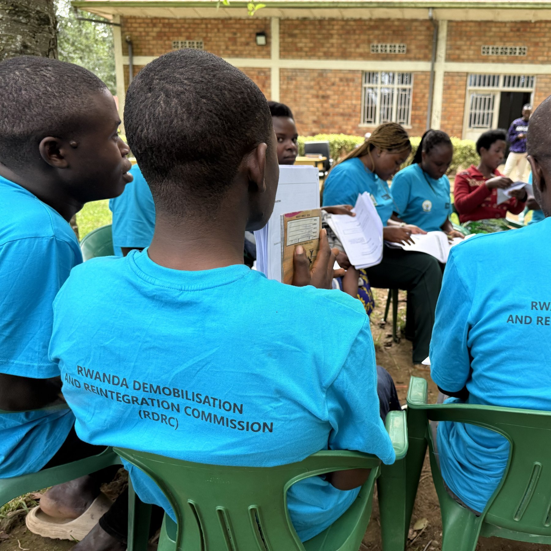 L'image montre un groupe de personnes assises à l'extérieur. Elles portent toutes des t-shirts bleu clair avec le texte "RWANDA DEMOBILISATION AND REINTEGRATION COMMISSION (RDRC)" imprimé au dos. On peut apercevoir des chaises en plastique vertes sur lesquelles elles sont assises. À l'arrière-plan, plusieurs personnes sont en train de discuter, et un bâtiment en briques apparaît légèrement flou derrière eux. L'ambiance semble conviviale et engagée, avec des participants qui semblent suivre une activité ou une formation en lien avec leur programme.