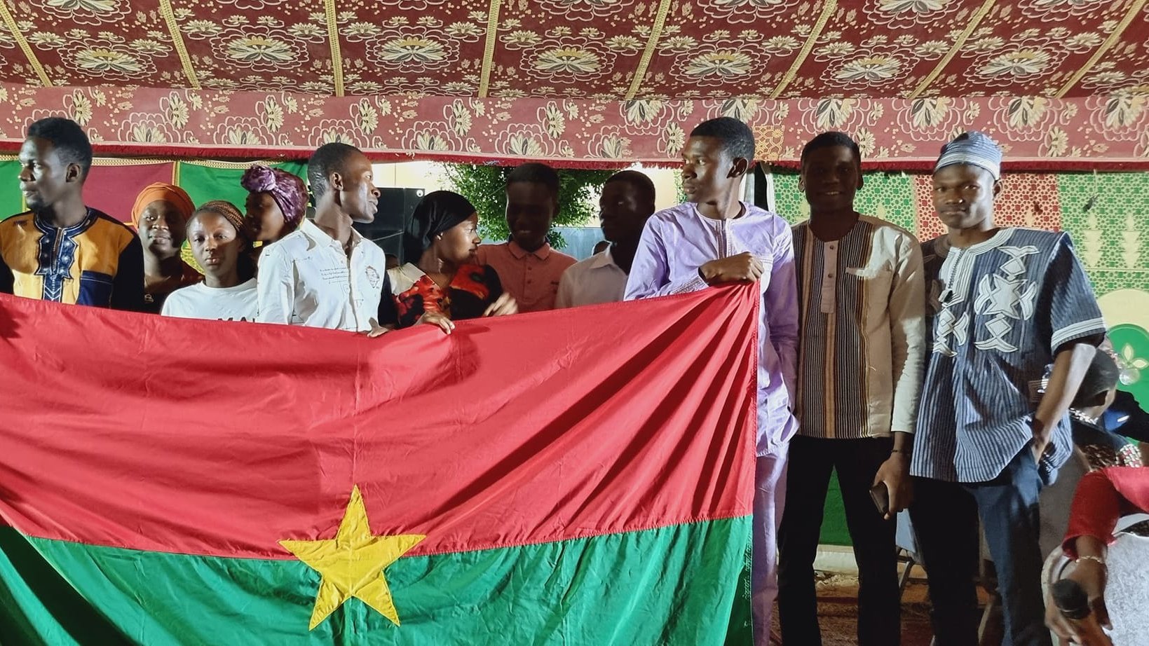 The image depicts a group of young people gathered together, holding a large flag of Burkina Faso, which features a red field with a green stripe at the bottom and a yellow star in the center. The setting appears to be festive, possibly during a cultural or community celebration, with decorative elements in the background. The individuals are wearing a mix of traditional and modern clothing, smiling and posing together, suggesting a sense of unity and pride in their national identity.
