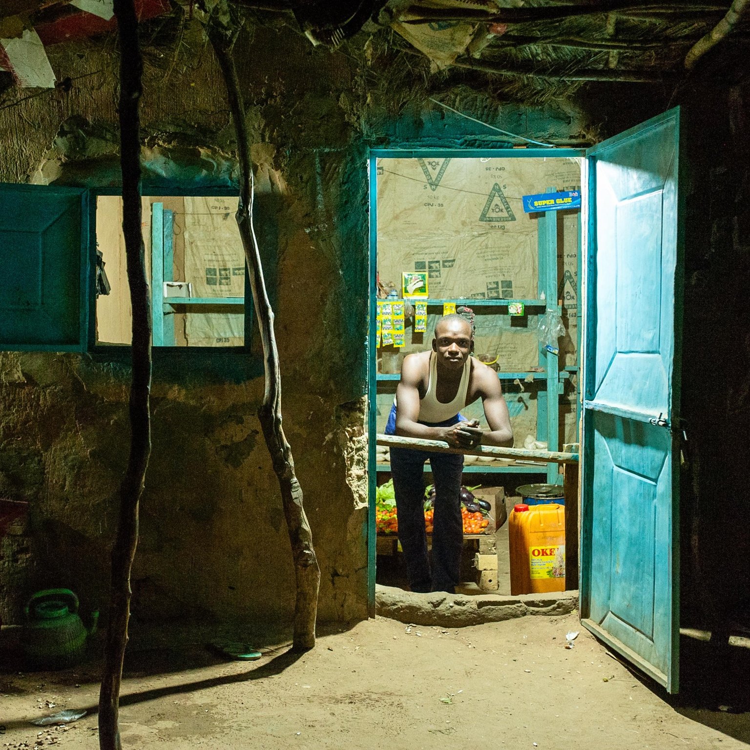 L'image montre l'entrée d'une petite boutique, éclairée dans l'obscurité. La porte est ouverte, révélant un intérieur modeste. On aperçoit un homme qui se tient derrière un comptoir, son regard tourné vers l'extérieur. Il porte un débardeur et semble attentif, probablement en attente de clients. Les murs sont en terre, et des papiers et affiches sont accrochés, ajoutant une touche de couleur à l'environnement. À l'avant, un banc en bois rouge est installé, et quelques bouteilles et contenants sont visibles à l'intérieur de la boutique. L'ambiance est chaleureuse malgré le contexte rural et simple.