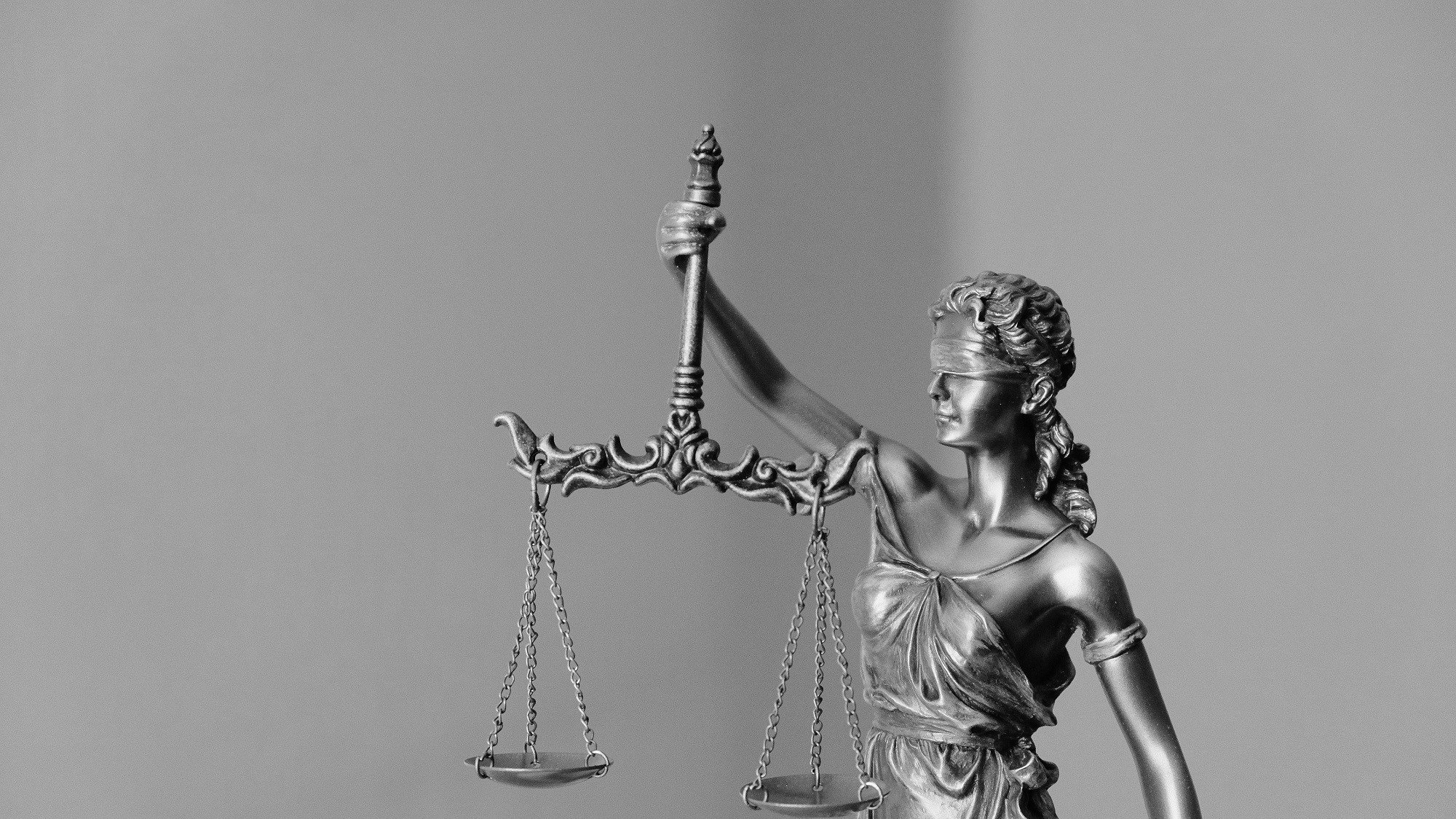L'image représente une statue allégorique de la justice. La figure féminine, vêtue d'une robe drapée, tient une balance dans une main, symbole d'équité et de mesure. Elle porte un bandeau sur les yeux, incarnant l'impartialité et l'objectivité. La statue est en métal et présente des détails raffinés, tels que des mèches de cheveux tressées. Le fond est simple et neutre, mettant en valeur la silhouette de la figure tout en accentuant son importance symbolique.