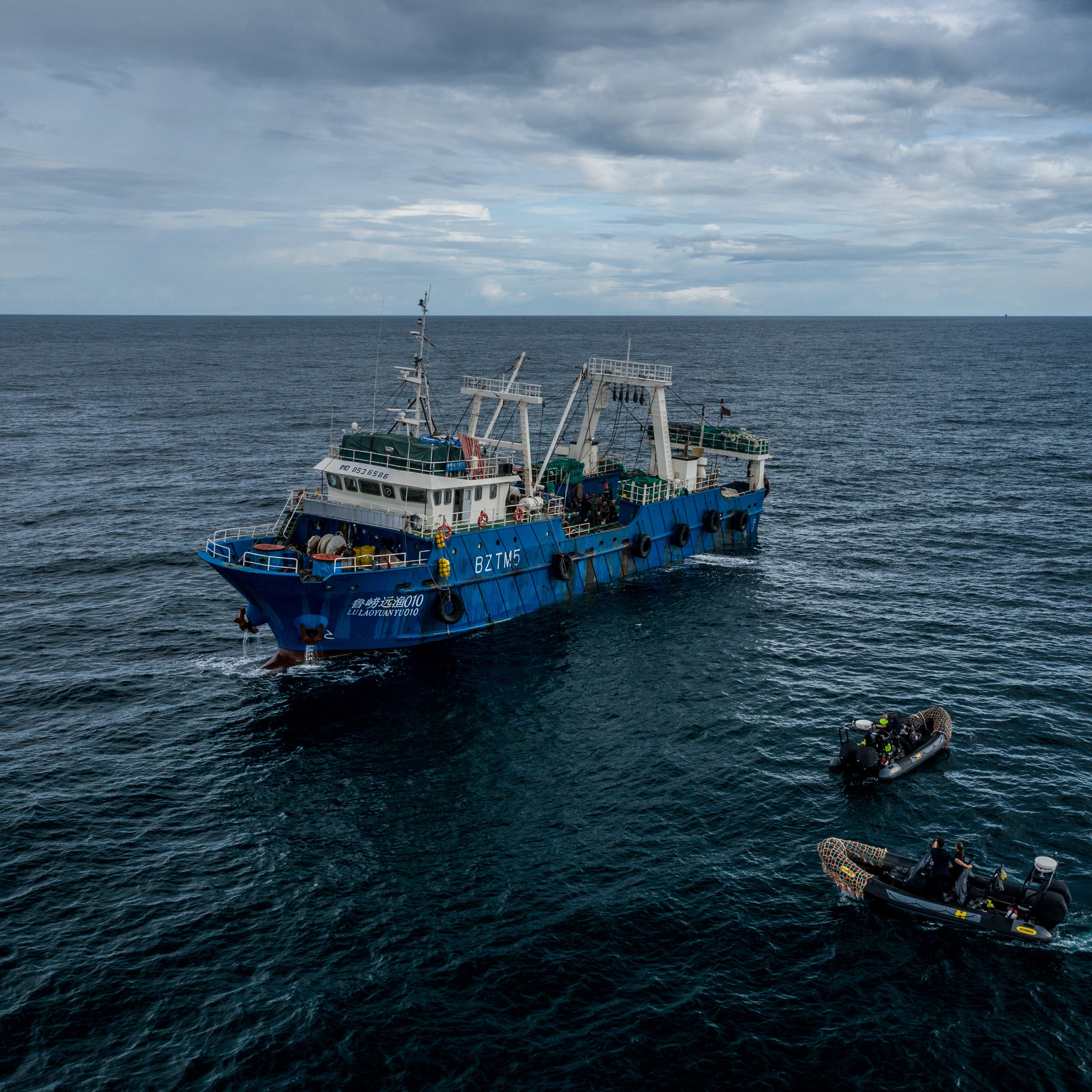 L'image montre un grand navire de pêche de couleur bleue, flottant au milieu de l'océan. Le bateau est en mouvement sur des eaux agitées, sous un ciel nuageux qui laisse présager une légère brise. À côté du navire principal, on peut voir deux petites embarcations, probablement des canots, qui sont en train de s'approcher. Les membres d'équipage, habillés en tenues de plongée, semblent occupés à des activités liées à la pêche. L'ambiance générale dégage une impression de travail maritime, avec le son des vagues et un léger souffle du vent.