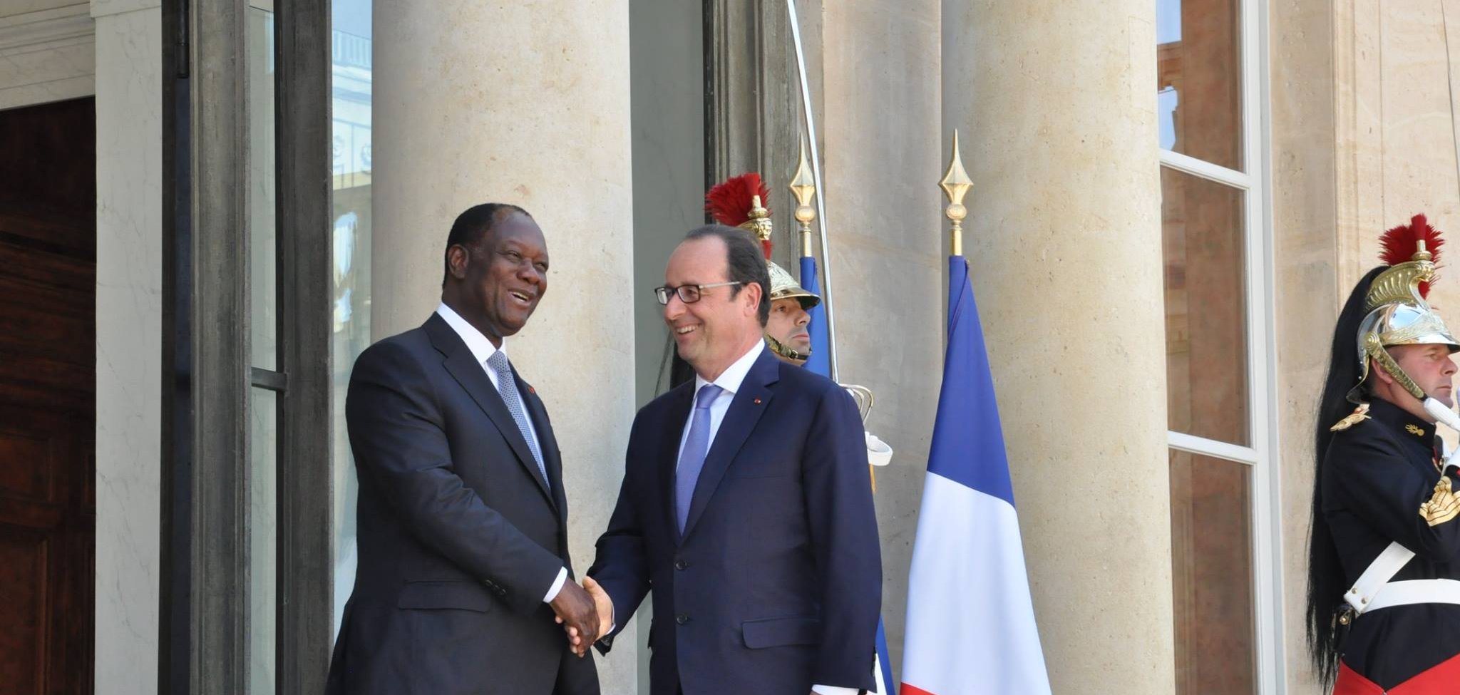 The image shows two men shaking hands outside a large building, likely a government or official residence. One man is wearing a dark suit and the other is dressed in a suit and tie, likely representing different nations. Behind them, there is a large flag of France, and guards in traditional uniforms stand watch. The setting suggests a formal meeting or diplomatic engagement.