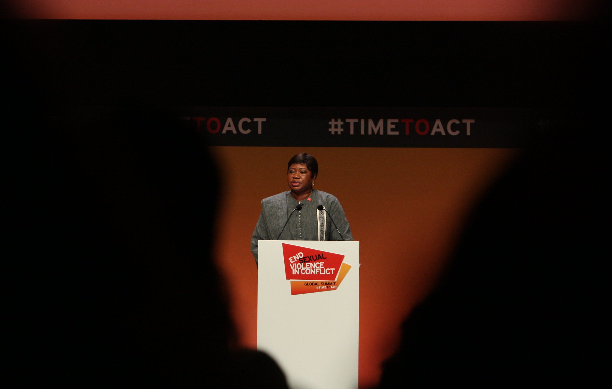 L'image montre une femme debout au micro, s'adressant à un public. Elle porte un vêtement formel et a une expression sérieuse. Le fond est principalement orange, avec des éléments graphiques en noir et rouge. Un panneau blanc devant elle affiche le texte "End Sexual Violence in Conflict" et "#TIMETOACT". La scène semble être un événement ou une conférence sur un sujet important, probablement lié à la lutte contre la violence sexuelle en contexte de conflit. Les silhouettes des personnes dans le public sont visibles en bas de l'image, ajoutant à l'ambiance de l'événement.