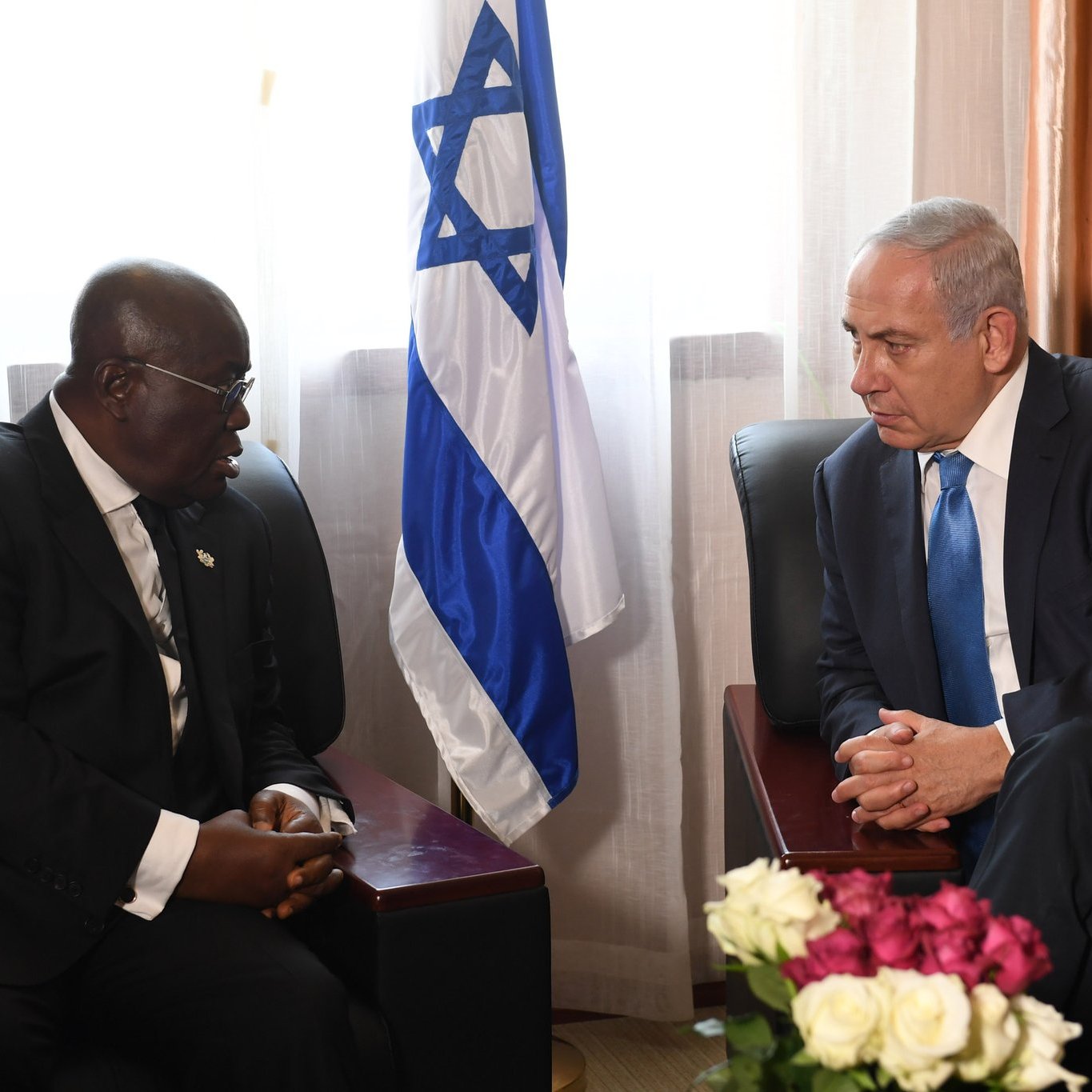 Dans cette image, deux hommes sont assis sur des canapés dans une pièce bien éclairée, entourés de rideaux lumineux. L'un des hommes, à gauche, porte un costume noir et des lunettes, tandis que l'autre, à droite, porte un costume clair et une cravate bleue. Entre eux, on peut voir une petite table avec un bouquet de fleurs aux teintes variées, principalement des roses blanches et des roses. En arrière-plan, se dresse un drapeau israélien, indiquant possiblement un contexte diplomatique. Les deux hommes semblent engagés dans une conversation sérieuse, chacun regardant l'autre avec attention.