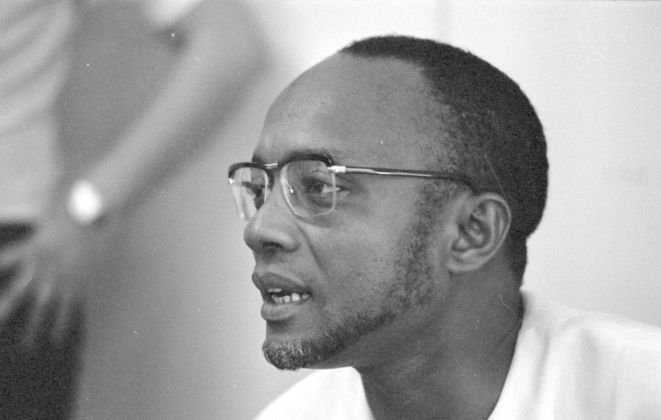 L'image représente un homme portant des lunettes à monture fine. Il a un visage allongé avec une petite barbe bien taillée. Ses cheveux sont courts et légèrement bouclés. L'homme semble concentré et engagé dans une conversation. En arrière-plan, on peut distinguer une silhouette floue d'une autre personne, suggérant qu'il se trouve dans un environnement social ou professionnel. L'éclairage est doux, ce qui met en valeur les traits de son visage.