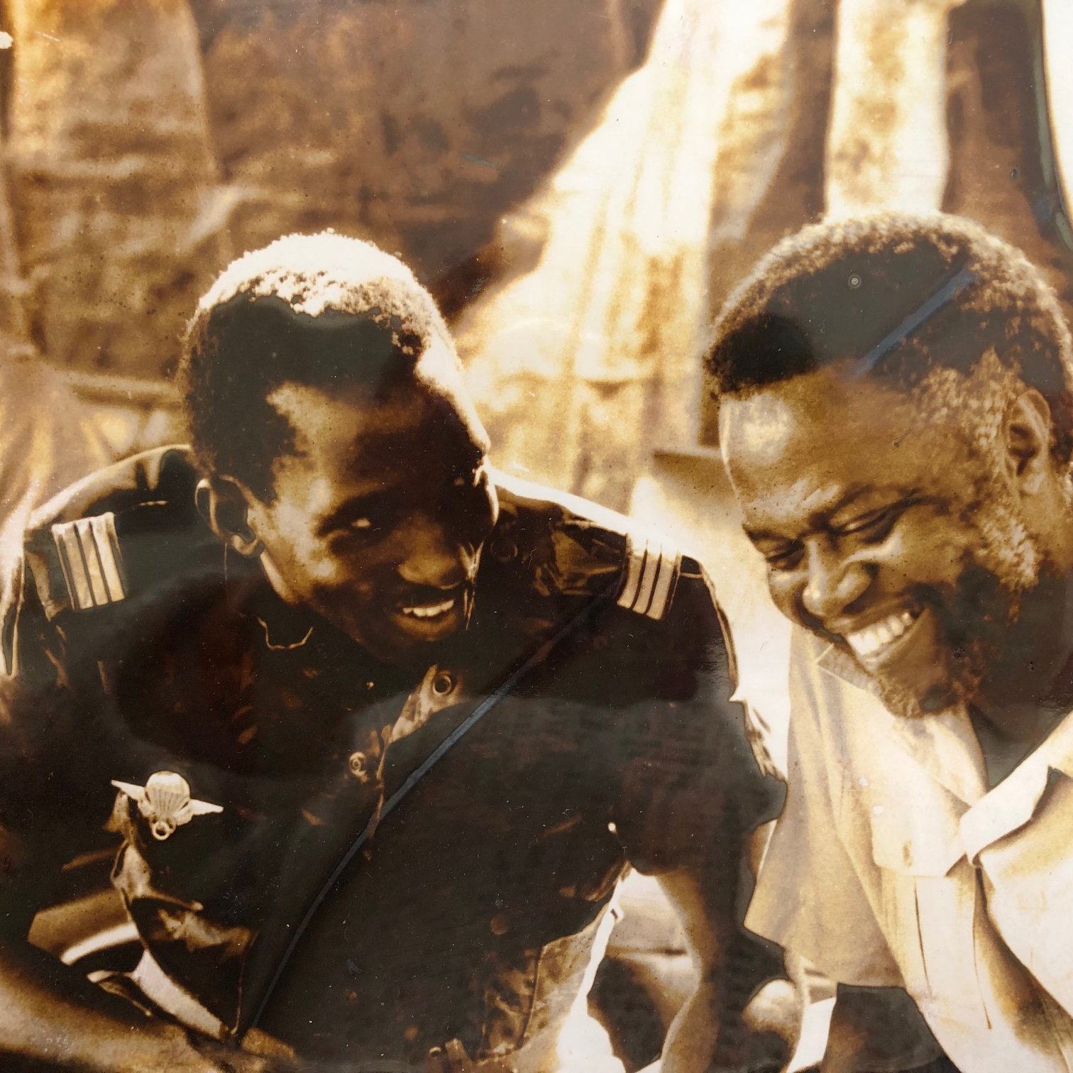 L'image présente deux hommes assis côte à côte, souriant et en train de discuter. L'un d'eux porte un uniforme militaire, tandis que l'autre est habillé d'une chemise claire. Leurs visages expriment la joie et la camaraderie. En arrière-plan, on peut distinguer des silhouettes floues de personnes et de la végétation, ce qui suggère un environnement social dynamique. Les teintes de l'image sont sépia, ce qui lui donne un aspect vintage, évoquant une époque passée.