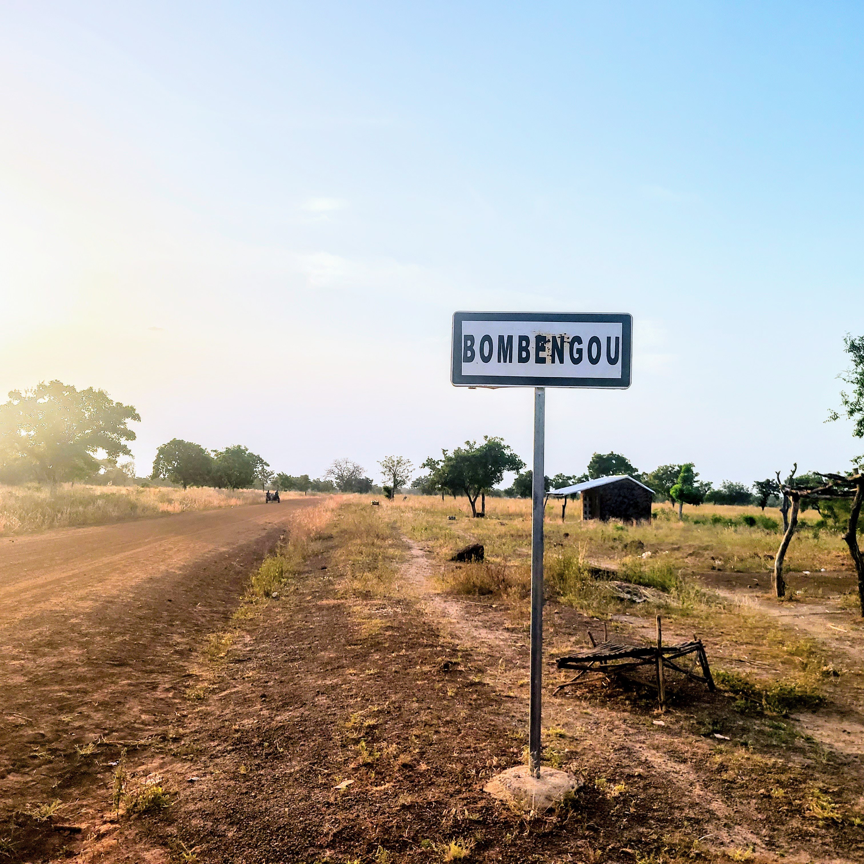 Cette image montre un panneau de signalisation indiquant le nom d'un village, "Bombengou", situé sur un chemin rural. Ce chemin est bordé de terre et de végétation, avec des arbres dispersés le long des côtés. La lumière du soleil crée une atmosphère douce et chaleureuse, suggérant un moment de la journée où la luminosité est agréable. En arrière-plan, on peut apercevoir une petite construction rudimentaire, peut-être une maison ou un abri. L'environnement semble calme et naturel, typique de la campagne.