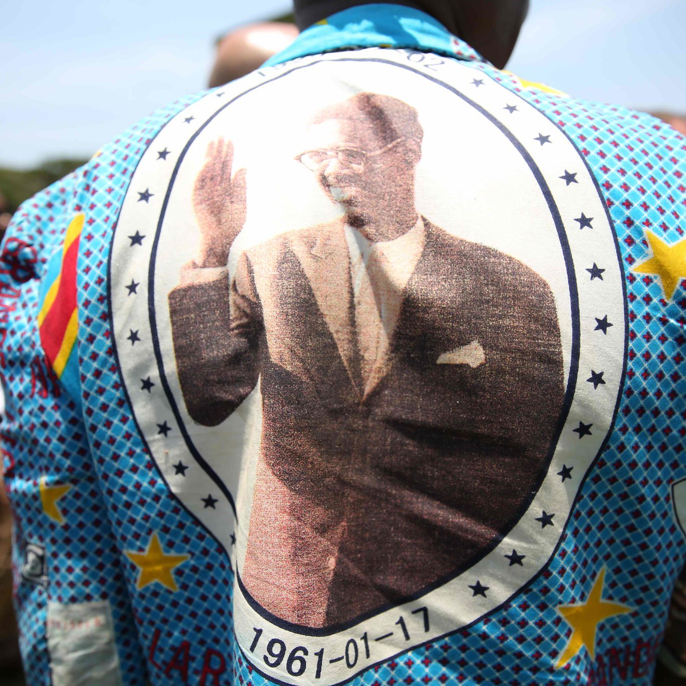 Cette image montre une personne portant un grand vêtement aux motifs colorés et vifs. Le dos du vêtement est orné d'une grande impression d'une personne en costume, levant la main dans un geste de salut. Autour de l'image, on peut voir des étoiles et des motifs géométriques. La date "1961-01-17" est inscrite, indiquant probablement une signification importante liée à l'image. En arrière-plan, on aperçoit un groupe de personnes, suggérant un événement ou une réunion en extérieur sous un ciel bleu.