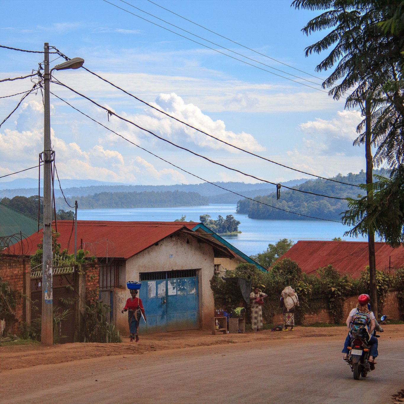 Voici une description de l'image : Vous vous trouvez sur une route située dans un paysage rural. À votre gauche, des maisons aux toits de tôle rouge et bleu se dressent, entourées de verdure luxuriante. Au loin, un lac scintille sous la lumière du soleil, reflétant les nuages blancs dans un ciel bleu. Certains habitants se déplacent, l'un d'eux portant une charge sur sa tête, tandis qu'un motard passe sur la route. Des poteaux électriques bordent la rue, ajoutant une touche de modernité à ce cadre naturel paisible. Le son lointain de l'eau et le chant des oiseaux peuvent également être présents dans cette ambiance tranquille.