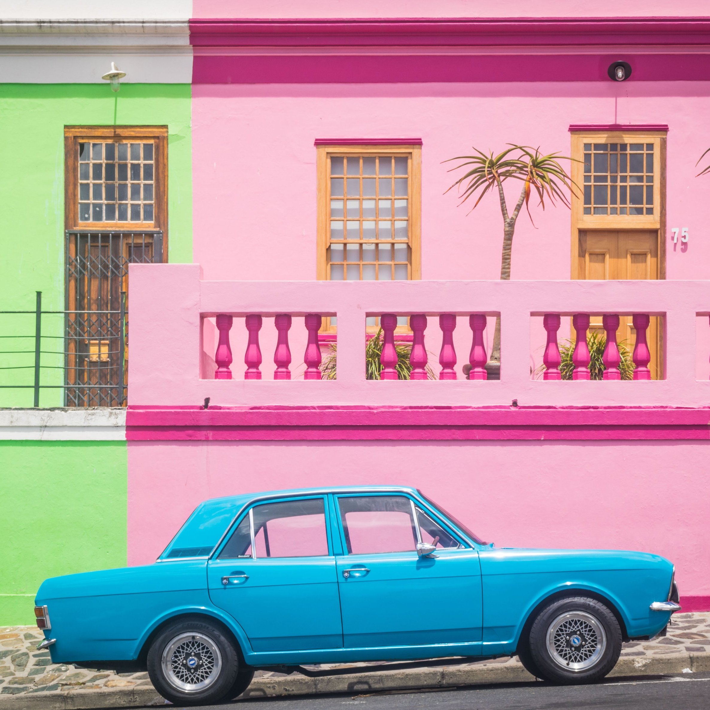 L'image présente une scène vibrante et colorée d'une rue typique. À gauche, un bâtiment se dresse en vert clair, avec des fenêtres en bois et un accès par escalier. À droite, un mur rose vif forme la façade d'une maison, ornée de deux palmiers qui ajoutent une touche tropicale. Entre les deux bâtiments, une voiture de couleur bleu clair est garée, se détachant joliment sur le fond coloré. Le sol est pavé, et l'ambiance générale de la scène est joyeuse et accueillante, évoquant une atmosphère estivale et ensoleillée.