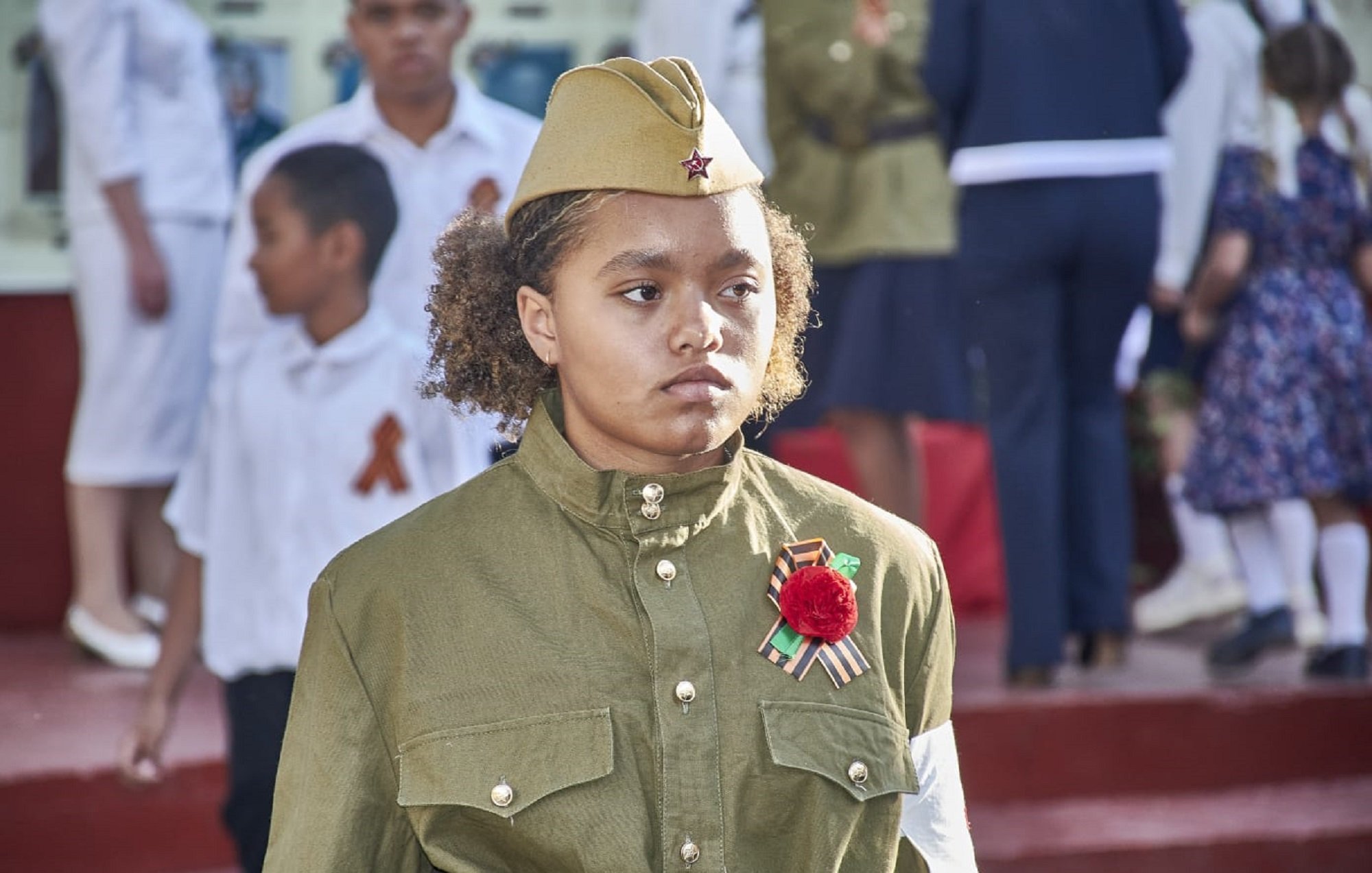 L'image montre une jeune fille portant un uniforme militaire, qui est de couleur olive et a des ornements spécifiques. Elle porte un chapeau similaire, typique des uniformes militaires. Sur sa poitrine, elle affiche une décoration, probablement une médaille ou un insigne, ainsi qu'une fleur rouge. En arrière-plan, on aperçoit d'autres personnes, dont des enfants en vêtements blancs et d'autres habits festifs, suggérant une célébration ou un événement commémoratif. L'expression de la jeune fille semble sérieuse, soulignant l'importance du moment. Le cadre est marqué par une ambiance solennelle, car plusieurs personnes semblent participer à une cérémonie.