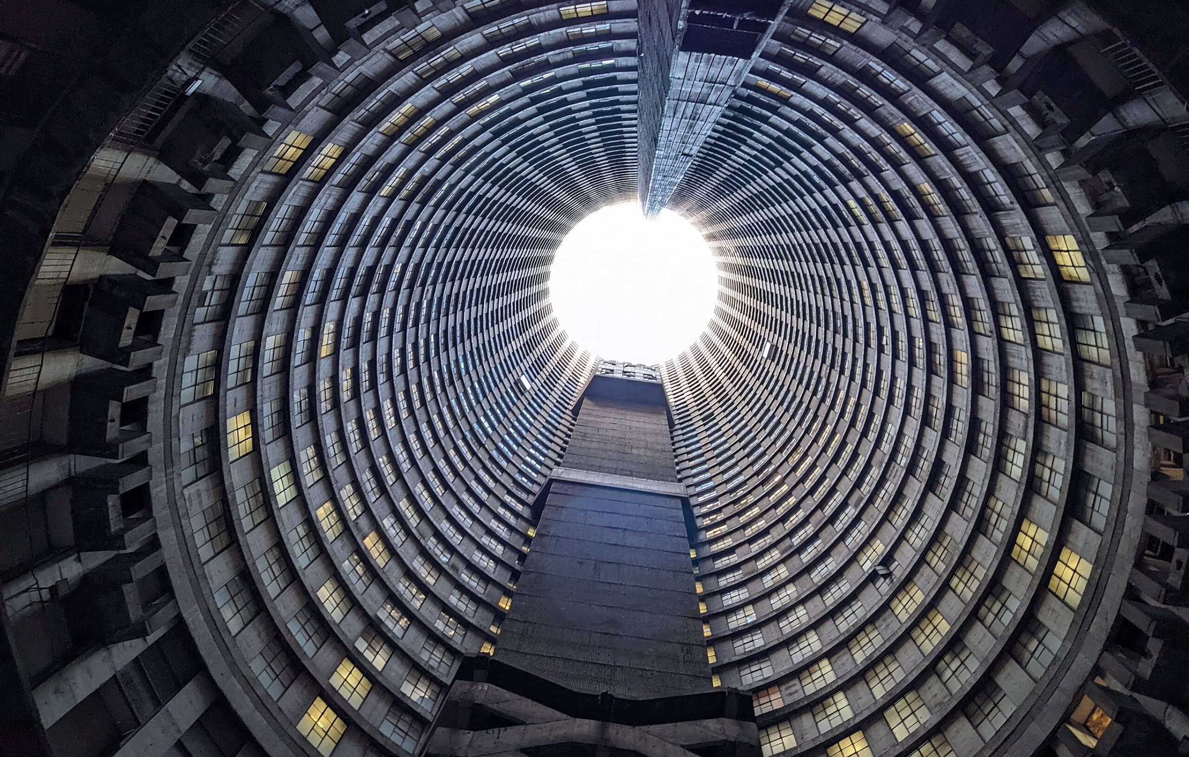 Imaginez vous tenir au centre d'une immense structure circulaire. La vue se dirige vers le haut, où un puits de lumière éclaire l'espace depuis l'ouverture au sommet. Les parois de ce bâtiment sont composées de béton et forment des motifs en spirale, créant une impression de profondeur et de hauteur. Les fenêtres qui bordent les étages, bien que nombreuses, laissent passer une lumière diffuse qui ajoute une sensation d'apaisement. Le sol est plat et peut sembler rugueux, contrastant avec la douceur de la lumière qui découpe le vide en haut. L'atmosphère est à la fois vide et pleine de possibilité, un mélange d'architecture industrielle et de nature, le tout se rejoignant dans ce grand espace ouvert.