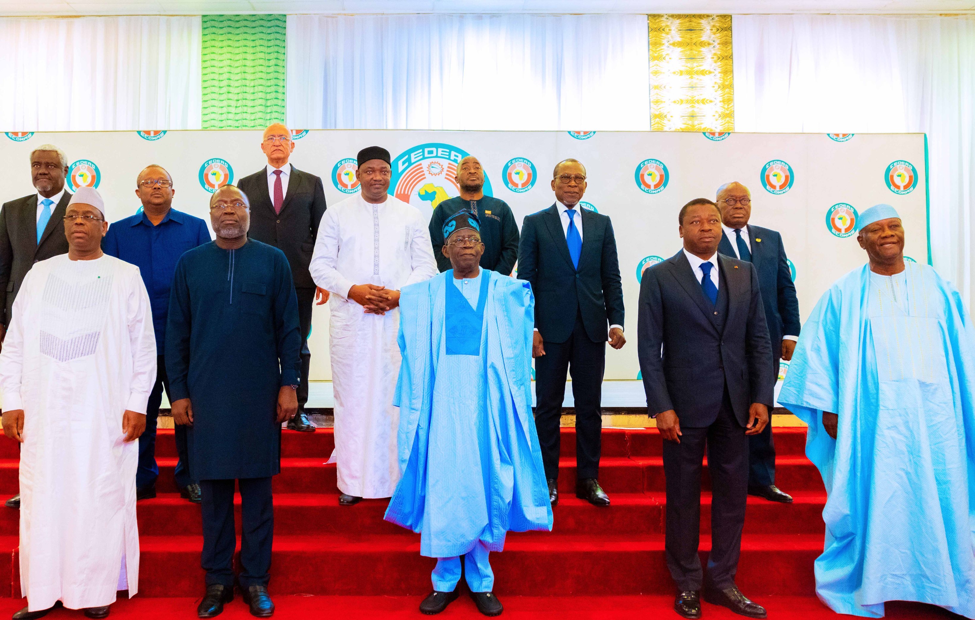 L'image montre un groupe de plusieurs hommes posant ensemble sur une scène. Ils sont vêtus de costumes formels et de tenues traditionnelles. La scène est décorée avec un fond de couleurs vives, comprenant un motif de logo sur la toile blanche. Les hommes se tiennent sur une estrade de couleur rouge, formant une sorte de demi-cercle. Certains portent des vêtements à motif et d'autres des costumes classiques, reflétant une diversité culturelle. L'atmosphère semble officielle, suggérant un événement important ou une réunion.