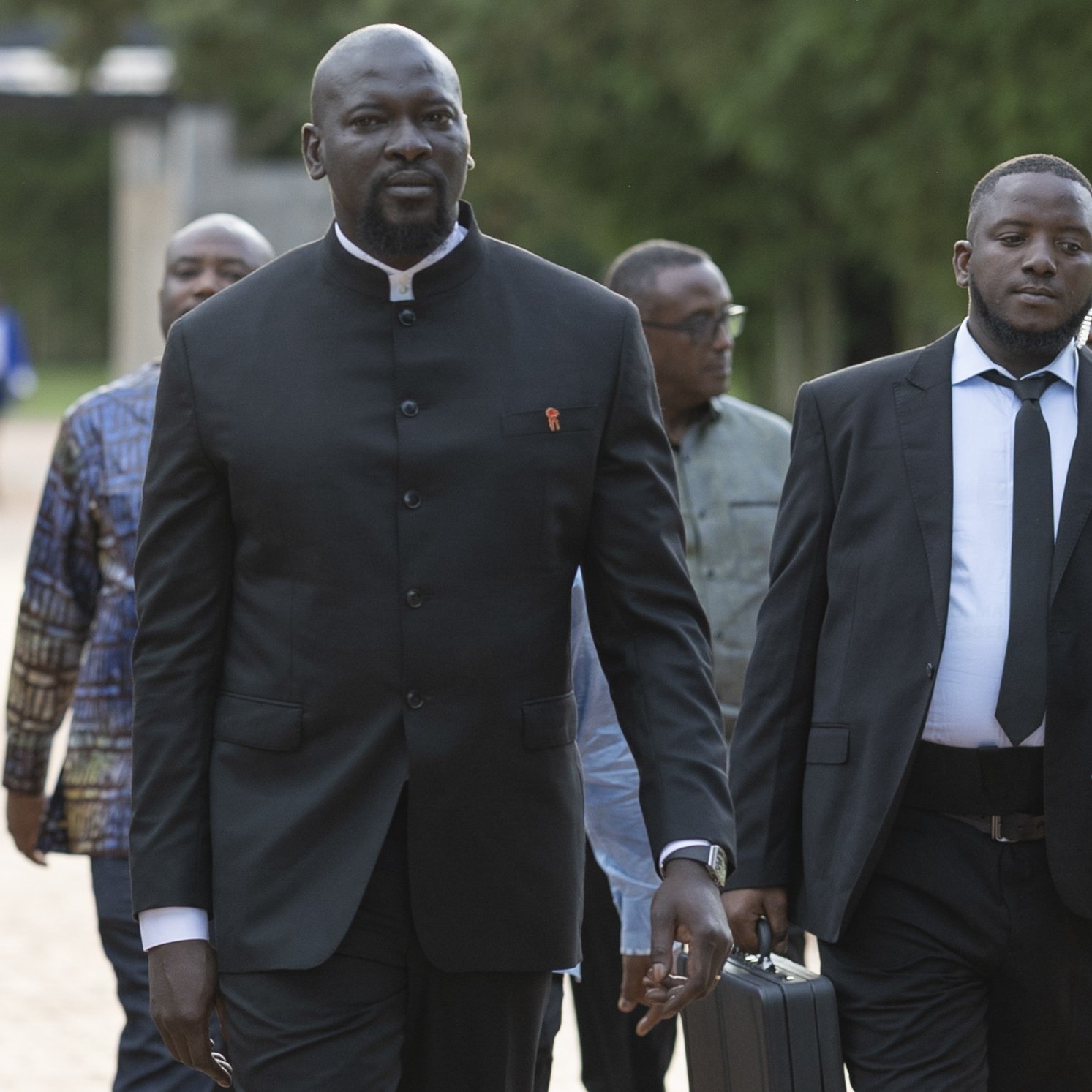 L'image montre un groupe d'hommes marchant ensemble sur un chemin dans un environnement verdoyant. Au premier plan, un homme grand et imposant porte un costume noir avec un col Mao, tandis qu'un autre homme à côté de lui est en costume noir et cravate. Ils ont l'air sérieux et concentrés, comme s'ils se rendaient à un événement important. En arrière-plan, d'autres hommes sont visibles, avec des tenues variées, ce qui suggère une atmosphère de réunion ou de conférence. Le paysage est paisible, avec des arbres et une lumière douce, ajoutant à l'ambiance formelle de la scène.