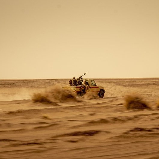 L'image montre un véhicule tout-terrain, probablement un pick-up, qui avance rapidement à travers un désert. La poussière s'élève autour de lui, créant une ambiance de mouvement et de vitesse. À l'arrière du véhicule, on aperçoit des silhouettes de personnes, probablement des passagers, qui semblent être équipés pour un environnement hostile. Le paysage est vaste et aride, avec des dunes de sable et des buissons résistant aux conditions sèches. Le ciel est clair mais teinté d'une couleur jaunâtre, suggérant une chaleur intense. L'ensemble donne une impression de solitude et d'aventure dans un milieu difficile.