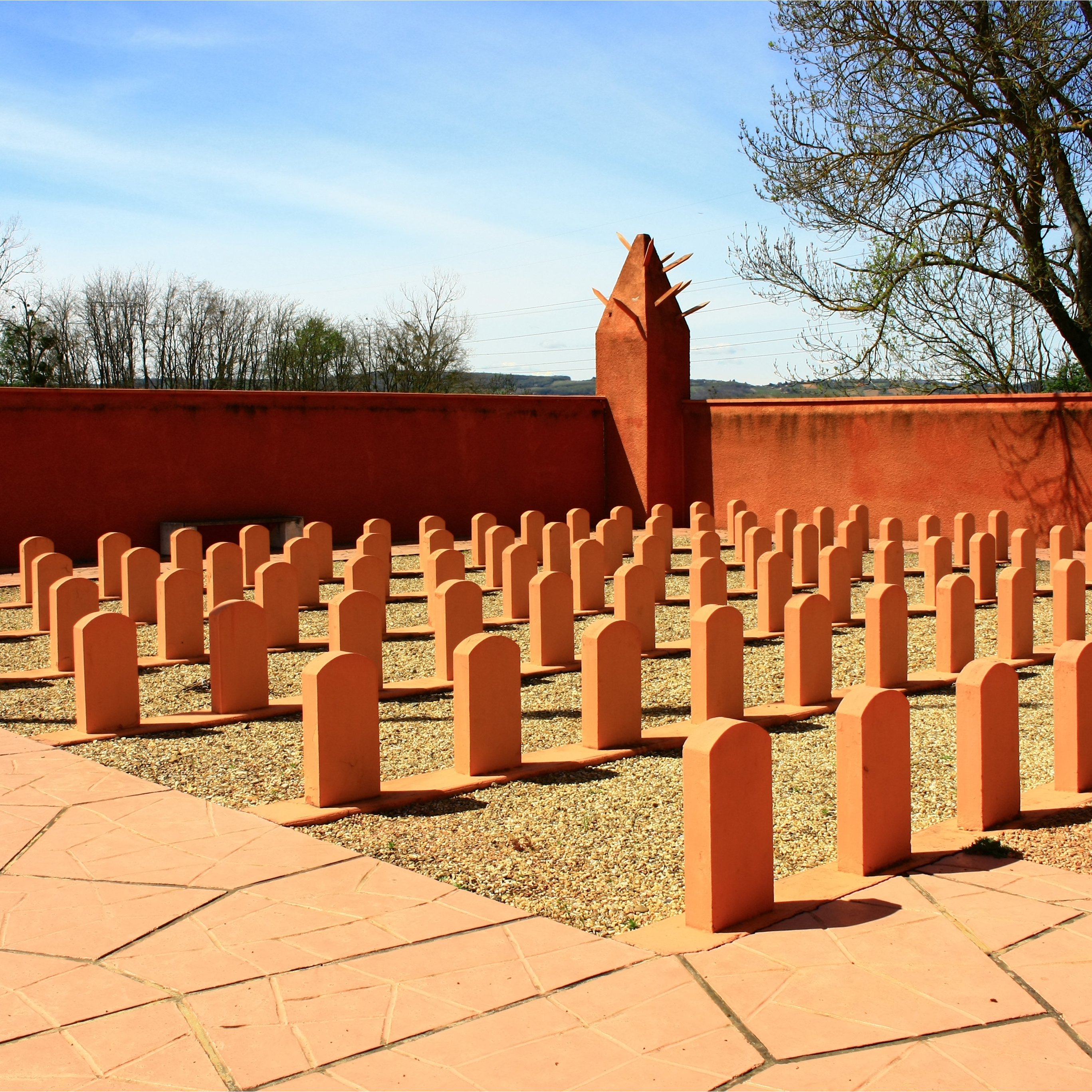 L'image montre un espace extérieur bien entretenu, avec plusieurs bornes ou stèles de forme allongée disposées en rangées sur un sol en gravier. Le sol est également pavé en terrasse, et il y a un mur de couleur beige orangé qui entoure la zone. En arrière-plan, des arbres dénudés se dressent contre un ciel bleu clair avec quelques nuages. L'ensemble dégage une atmosphère de sérénité et de réflexion.