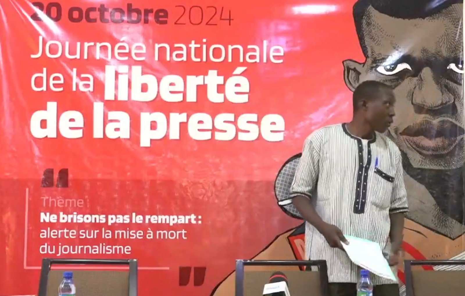 L'image montre un événement lié à la Journée nationale de la liberté de la presse, qui se déroule le 20 octobre 2024. En arrière-plan, un grand panneau rouge présente le thème de la journée : "Ne brisons pas le rempart : alerte sur la mise à mort du journalisme". Au premier plan, un homme debout s'apprête à s'adresser à l'audience. Il porte une chemise à rayures noires et blanches et se tient près d'une table équipée de plusieurs microphones et de bouteilles d'eau. L'ambiance est sérieuse et engageante, soulignant l'importance de la liberté d'expression.