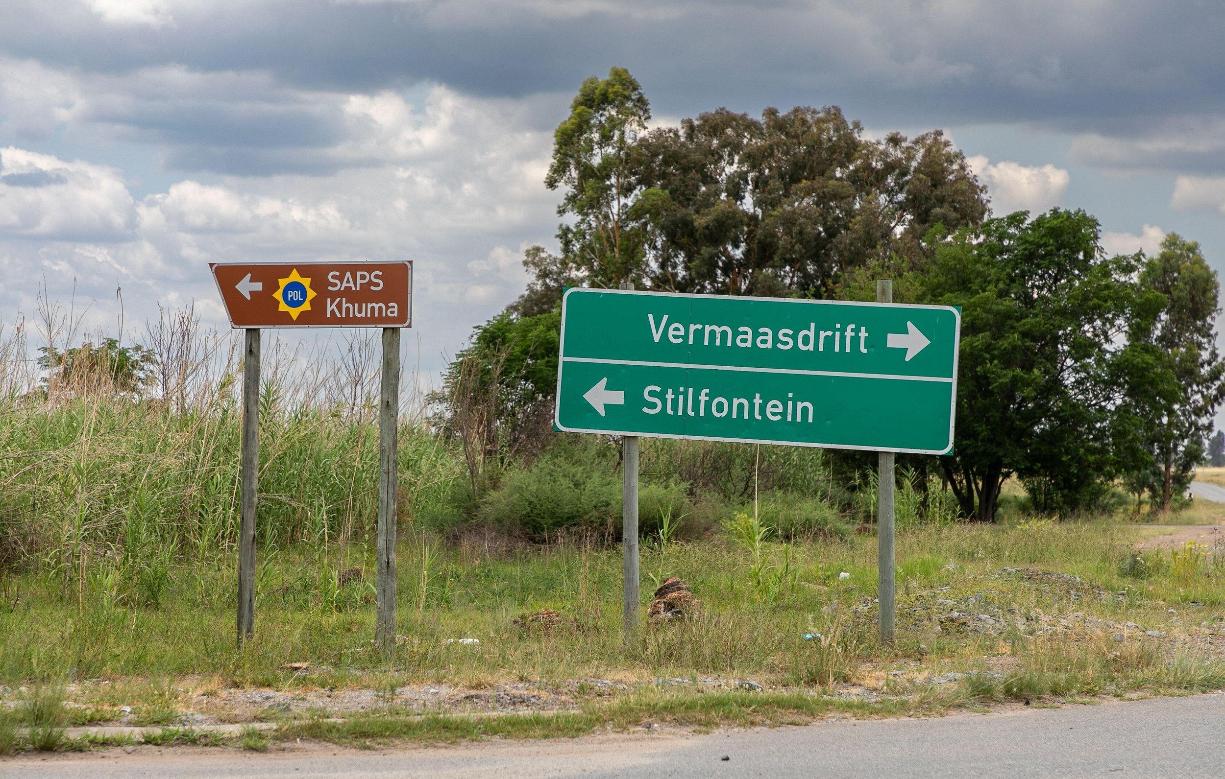 Cette image montre un carrefour routier en extérieur. À gauche, un panneau marron avec un symbole de police indique "SAPS Khuma", suggérant la présence d'un poste de police local. À droite, un panneau vert montre deux flèches directionnelles : une vers "Vermaasdrift" et l'autre vers "Stilfontein". L'environnement est caractérisé par une végétation verte et dense, avec quelques arbres en arrière-plan et un ciel nuageux, créant une atmosphère calme et naturelle. Le sol est asphalté, donnant l'impression d'une route peu fréquentée.