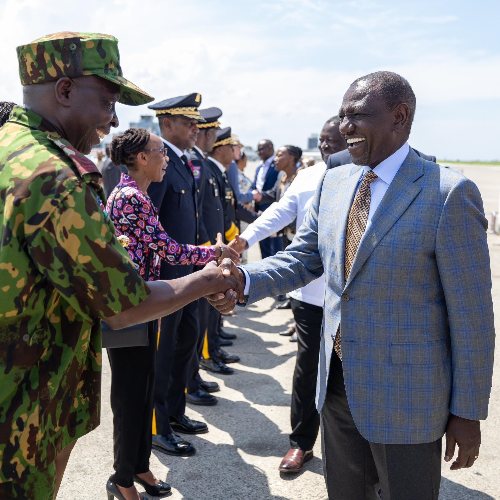The image shows a formal scene at an airport where a group of people is gathered, likely to welcome a dignitary or a visitor. In the foreground, two men are shaking hands; one is dressed in military camouflage, and the other is in a formal suit. Around them, there are officials in uniform and business attire lined up, indicating a significant or ceremonial occasion. The setting seems to be sunny, contributing to a positive atmosphere.