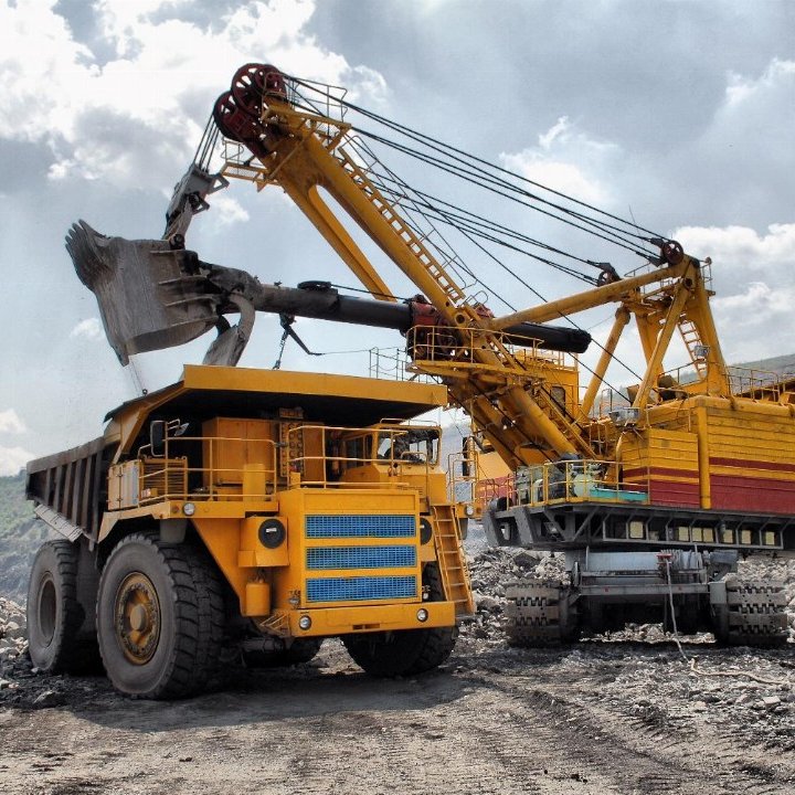 L'image présente un site d'extraction minière. Au premier plan, un gros camion de transport de couleur jaune, avec des roues massives, est chargé de matériaux. À côté, une grande pelle mécanique jaune et rouge, dotée d'un bras articulé, a relevé une charge de terre ou de roches. Le paysage environnant est accidenté, avec des tas de gravier et des pentes abruptes. Le ciel est partiellement nuageux, laissant passer des rayons de lumière qui illuminent la scène, créant une ambiance industrielle dynamique. Des bruits de machines et d'activités minières peuvent être imaginés, rendant l'environnement à la fois impressionnant et robuste.