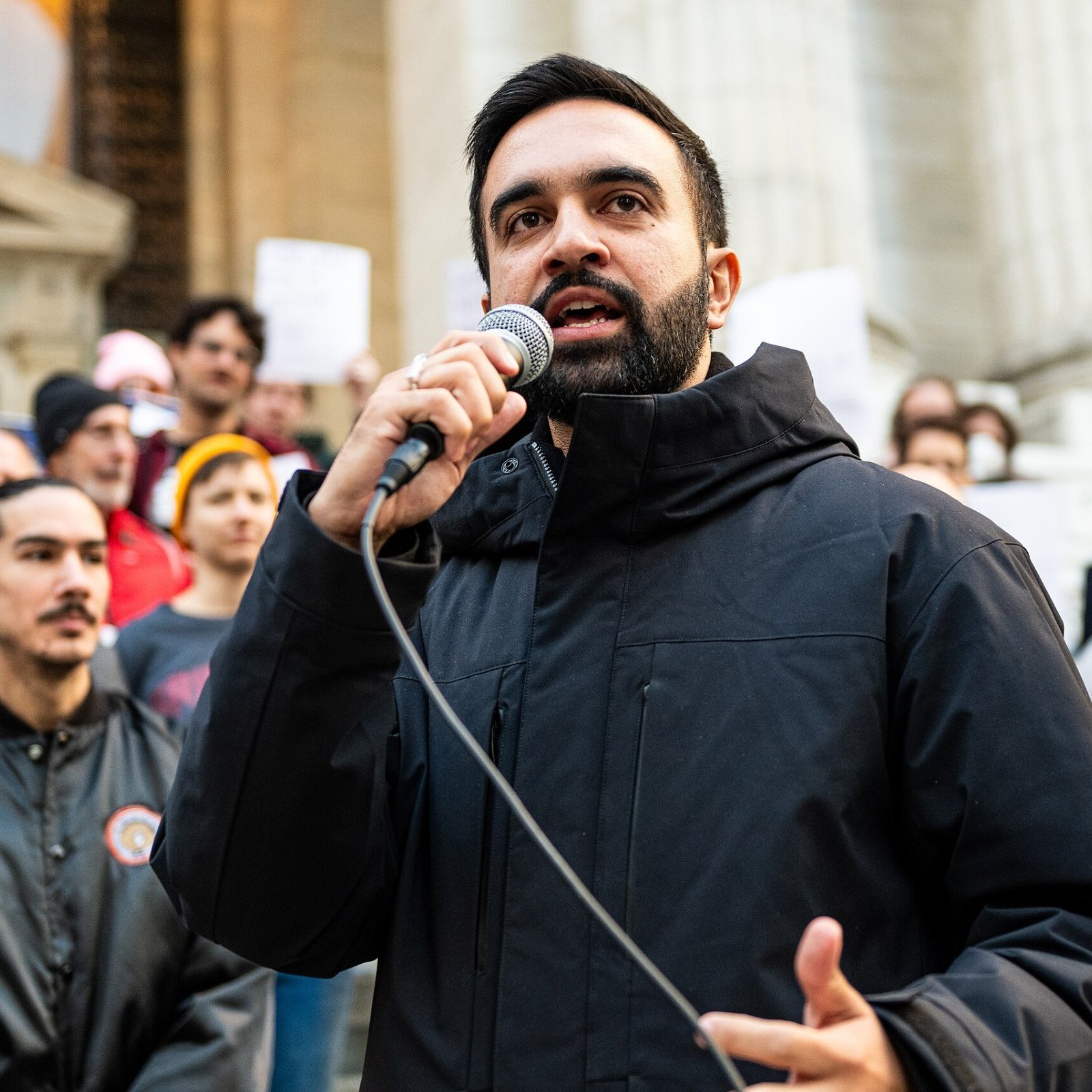 L'image montre un homme debout en avant-plan, tenant un microphone dans sa main et s'adressant à une foule. Il porte une veste noire et a une expression déterminée sur son visage. En arrière-plan, on aperçoit un groupe de personnes, certaines tenant des affiches. L'environnement semble être une place publique, avec des bâtiments importants en arrière-plan. L'atmosphère dégage une impression de mobilisation et d'engagement collectif.