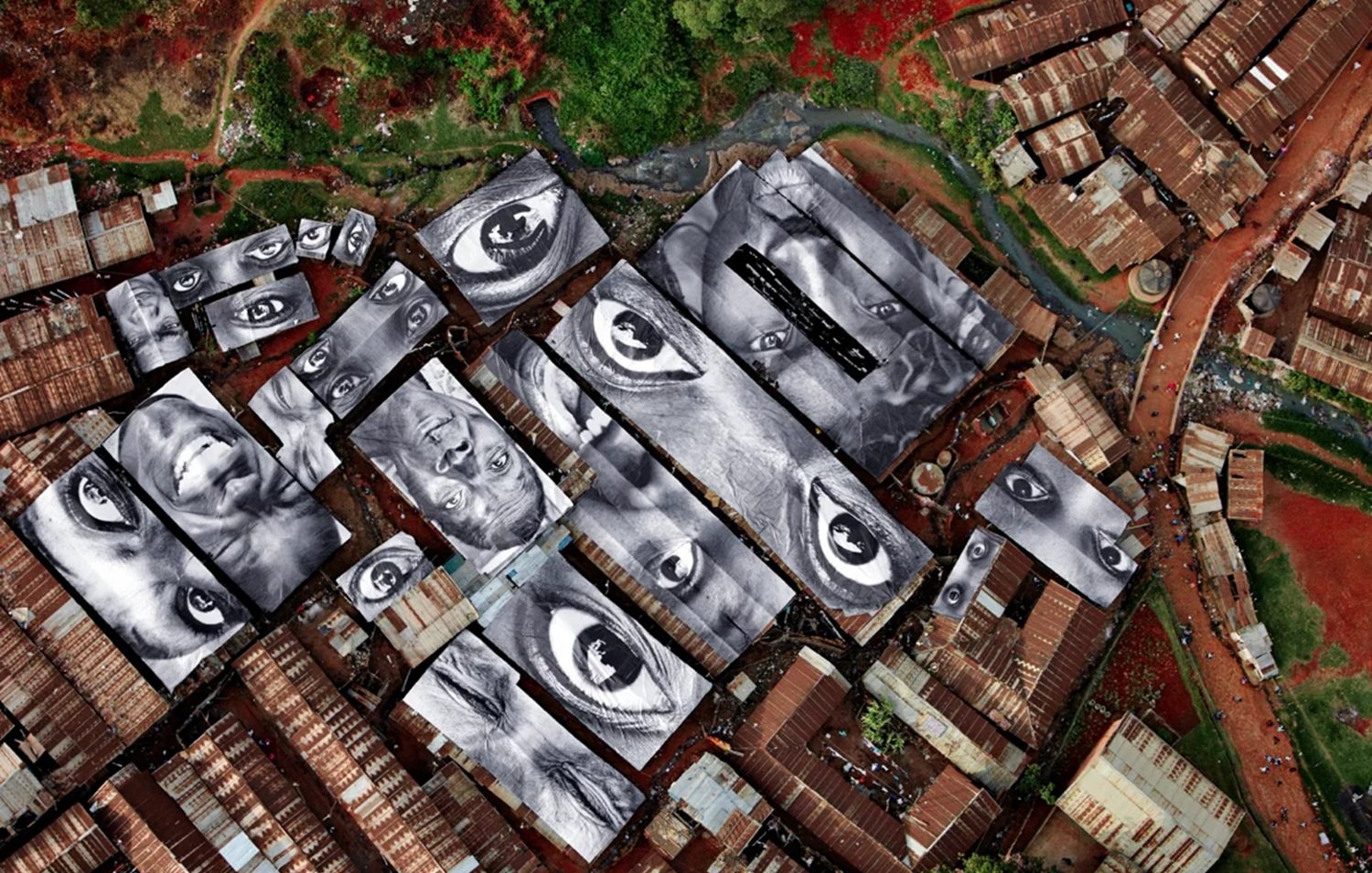 L'image représente une vue aérienne d'un bidonville, où les toits en tôle ondulée sont de différentes teintes, principalement marron. Sur ces toits, il y a de grandes œuvres d'art en noir et blanc illustrant des visages humains. Les yeux des portraits semblent fixer le spectateur, créant une impression d'intensité et de connexion. Entre les toits, on aperçoit des espaces plus clairs, peut-être des chemins ou des ruelles, et on peut deviner la verdure environnante qui ajoute une touche de contraste à l'environnement urbain. L'ensemble évoque à la fois la pauvreté du lieu et la richesse des émotions humaines capturées dans les portraits.