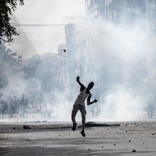 L'image dépeint une scène urbaine troublée, où la tension est palpable. Au premier plan, une silhouette humaine, probablement un homme, est en mouvement, lançant un projectile. Il semble néanmoins être entouré de foules, suggérant une situation tumultueuse. Des nuages de fumée épaisse s'élèvent dans l'air, créant une atmosphère de chaos, tandis que des débris jonchent le sol, témoignant d'une agitation récente. En arrière-plan, on peut deviner des bâtiments urbains flous, et la lumière est sombre, renforçant l'impression d'une émeute ou d'une protestation. Les sons et odeurs d'une ville agitée pourraient également accompagner cette image, avec des cris, des sirènes, ou l'odeur de fumée.