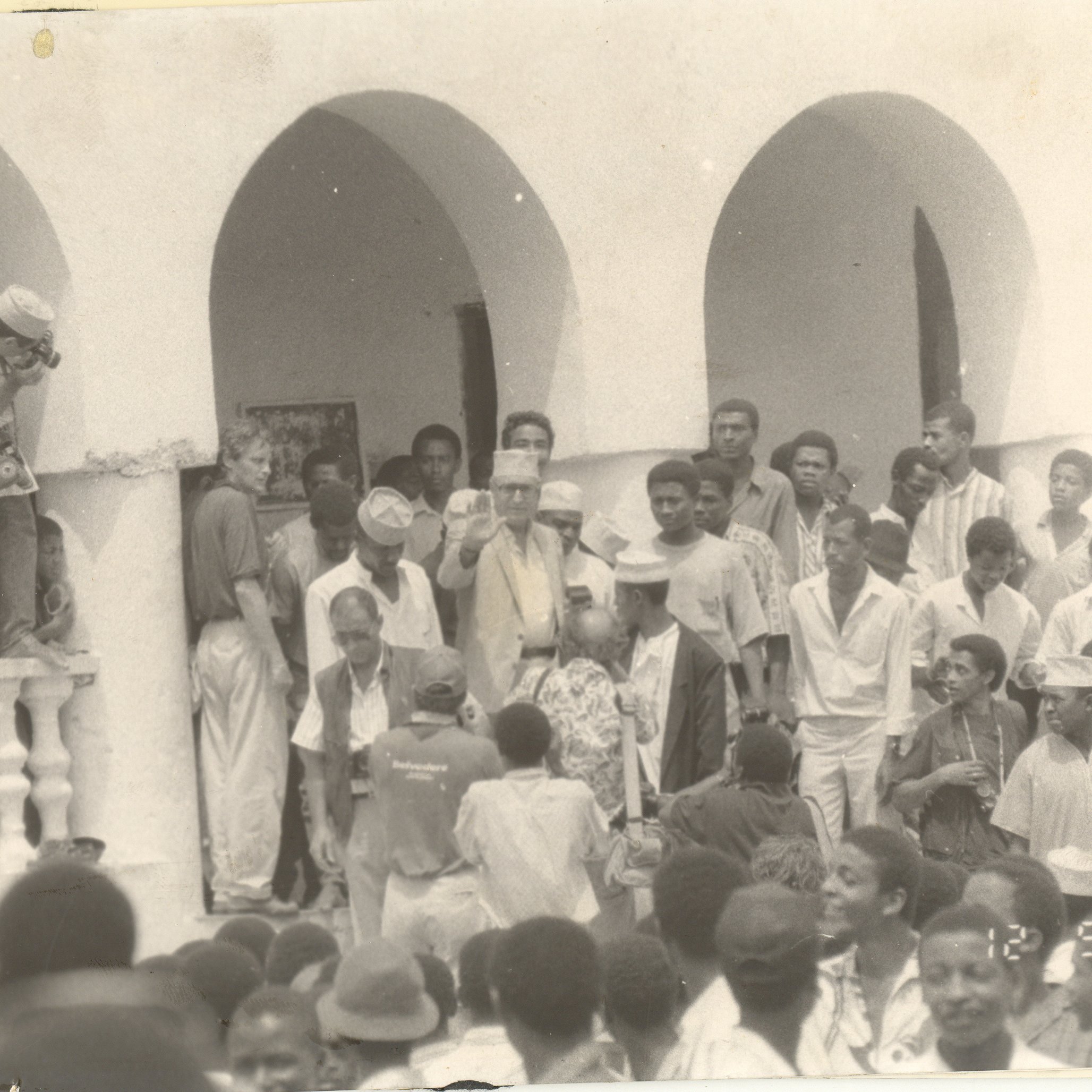 L'image représente une scène de rassemblement en extérieur, probablement lors d'un événement communautaire ou historique. Au premier plan, un groupe de personnes se regroupe autour d'une figure centrale, tandis qu'une autre personne, en haut à gauche, semble prendre une photo. Les participants portent des vêtements variés, certains avec des chapeaux. L'architecture en arrière-plan témoigne d'un style colonial, avec des arches et des balustrades. L'ambiance générale paraît vivante et joyeuse, suggérant un moment d'unité et de partage au sein d'une communauté.