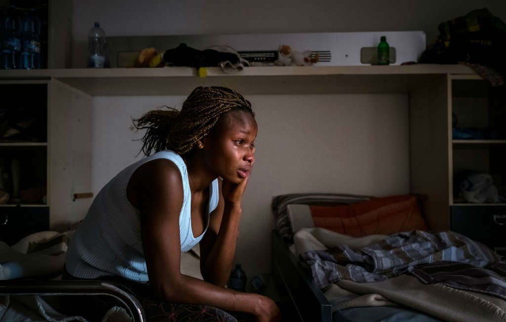 L'image dépeint une femme assise sur le bord d'un lit dans une pièce sobre. Elle a une coiffure tressée et porte un haut sans manches de couleur claire. Son expression est pensive, montrant une profonde réflexion ou peut-être de la tristesse. La lumière douce qui filtre à travers une fenêtre éclaire son visage, créant des ombres sur ses traits. La chambre semble être en désordre, avec des objets éparpillés et des draps non rangés. L'atmosphère de l'image évoque un sentiment de solitude et de contemplation.