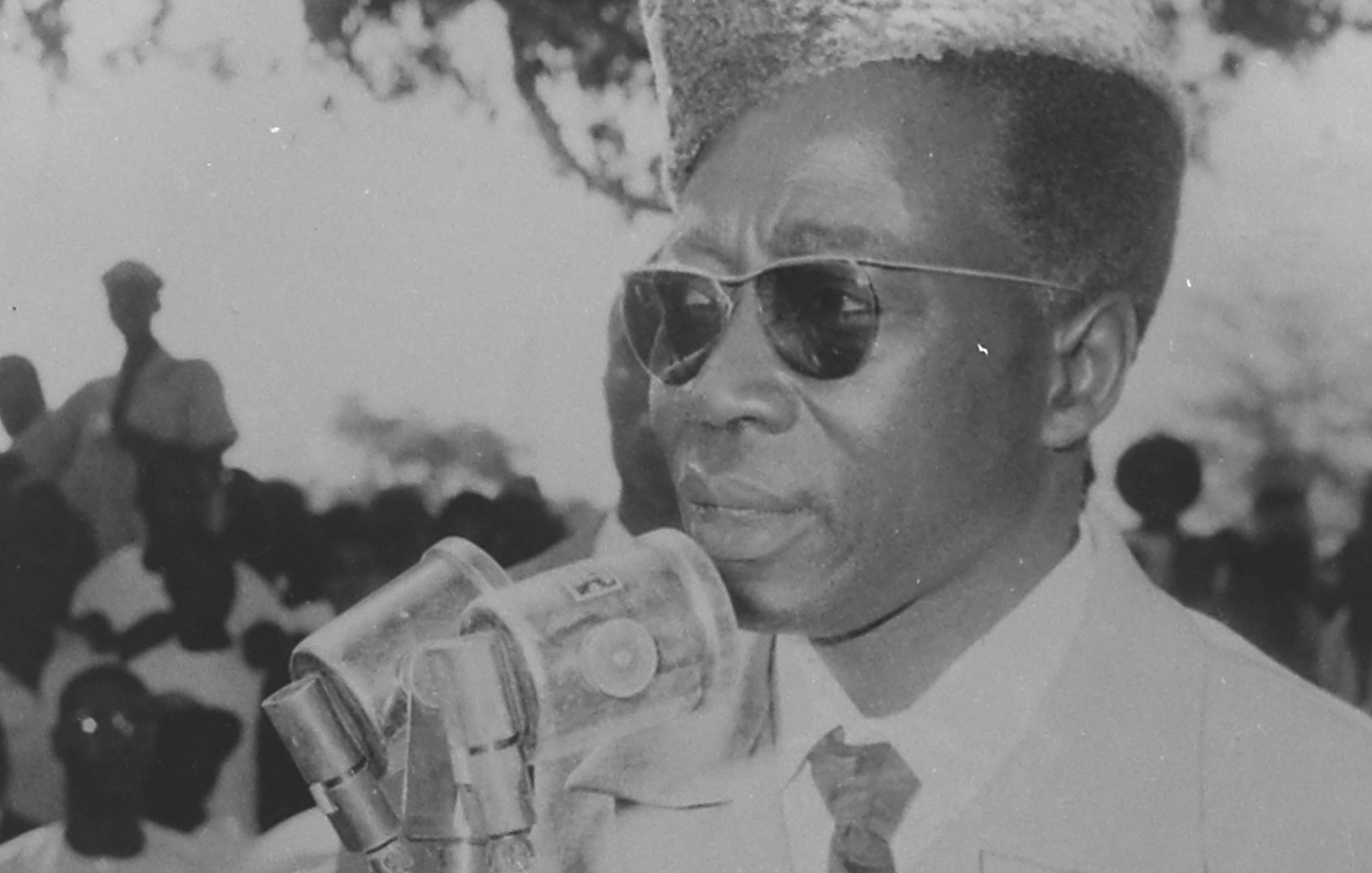 L'image montre un homme debout devant un microphone, s'exprimant clairement. Il porte des lunettes de soleil et a une coiffure distinctive, avec une coupe qui semble bien soignée. Il est vêtu d'un costume clair, ce qui ajoute à son allure sérieuse. En arrière-plan, on peut apercevoir une foule de personnes, dont certaines sont floues, semblant écouter attentivement. L'atmosphère de la scène évoque un moment important, peut-être un discours ou une déclaration publique.