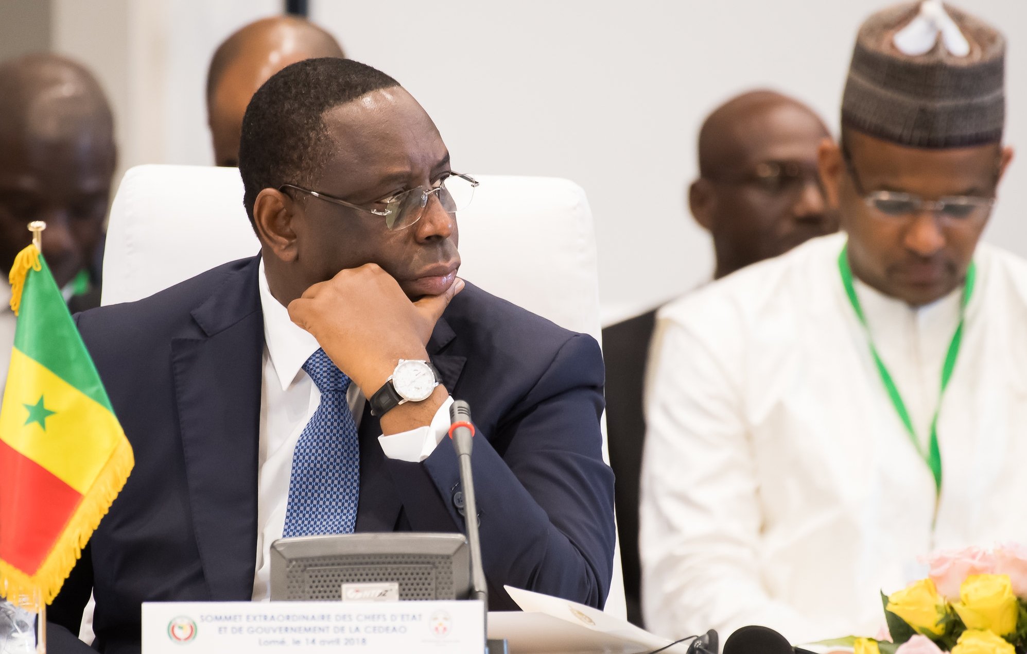 L'image montre un homme assis à une table dans un cadre formel. Il porte un costume sombre et un bracelet montre argenté. Son visage exprime une concentration sérieuse, alors qu'il regarde pensivement devant lui. À sa gauche, il y a un drapeau du Sénégal planté sur la table, indiquant son origine ou son rôle. En arrière-plan, d'autres personnes sont présentes, mais ne sont pas clairement visibles. L'ambiance de la scène suggère un événement officiel, peut-être une réunion ou une conférence de dirigeants.