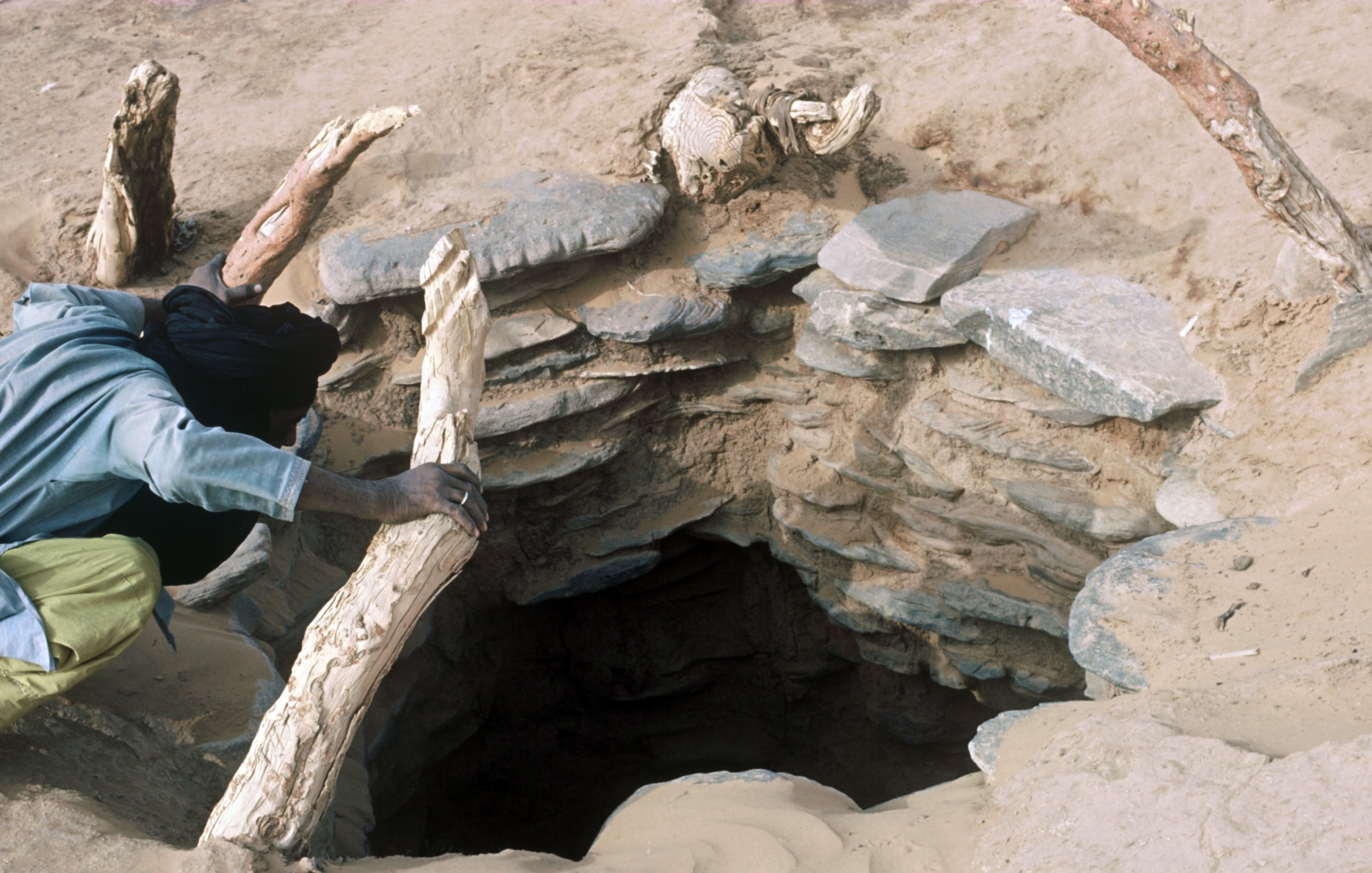 L'image montre un trou dans le sol, probablement un puits, entouré de pierres disposées de manière circulaire. À côté du trou, on aperçoit une personne, vêtue d'une chemise et d'un pantalon longs, qui semble s'approcher ou manipuler un morceau de bois. Le terrain est sablonneux, et des débris de bois sont visibles autour, suggérant une utilisation régulière de cet endroit. L'atmosphère semble aride, reflétant un environnement désertique.
