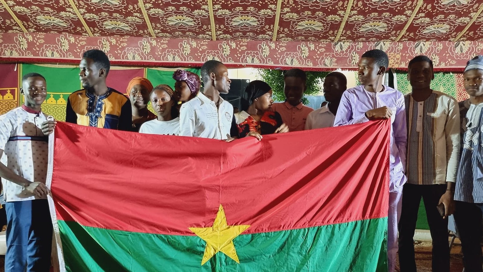 L'image présente un groupe de jeunes personnes rassemblées sous une tente décorée, avec des motifs colorés en arrière-plan. Au centre, ils tiennent une grande drapeau du Burkina Faso, qui est rouge avec une étoile verte au milieu. Les membres du groupe affichent des vêtements variés, certains portant des chemises traditionnelles, et semblent joyeux et solidaires. L'atmosphère est festive, suggérant un événement culturel ou une célébration.