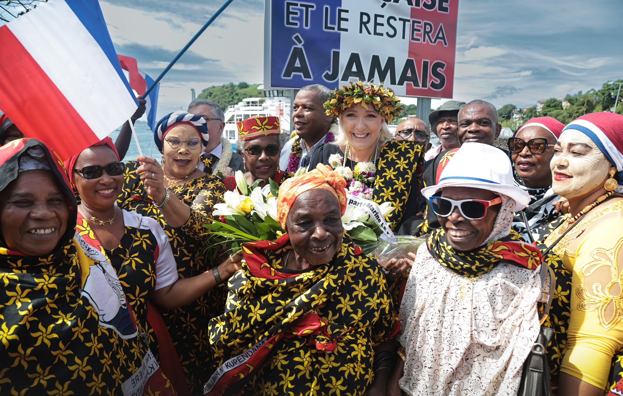 L'image représente une scène festive, probablement lors d'une célébration culturelle ou d'un événement politique. Au centre, plusieurs femmes et hommes portent des vêtements colorés, souvent ornés de motifs floraux jaunes sur fond noir. Certains participants tiennent des bouquets de fleurs tandis que d'autres brandissent des drapeaux français. Au fond, un grand panneau affiche un message qui semble souligner un attachement fort à la France, avec les mots "ET LE RESTERA À JAMAIS". L'atmosphère est joyeuse, avec des sourires et des expressions de célébration visible sur les visages des participants. Le ciel en arrière-plan est partiellement nuageux, ajoutant à l'ambiance vibrante de l'événement.