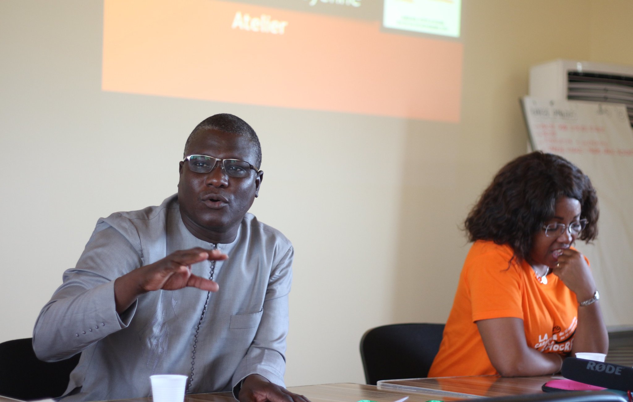 L'image montre un atelier où deux personnes sont assises à une table. À gauche, un homme, portant des lunettes et une tenue traditionnelle grise, gesticule avec une main tout en parlant. Son expression est engageante et dynamique, suggérant qu'il s'adresse à un groupe. À droite, une femme, habillée d'un t-shirt orange, écoute attentivement. Son visage montre une attitude attentive et intéressée. En arrière-plan, un écran est visible avec des textes, mais il est flou. L'atmosphère semble interactive et collaborative, indiquant un échange d'idées lors de l'atelier.