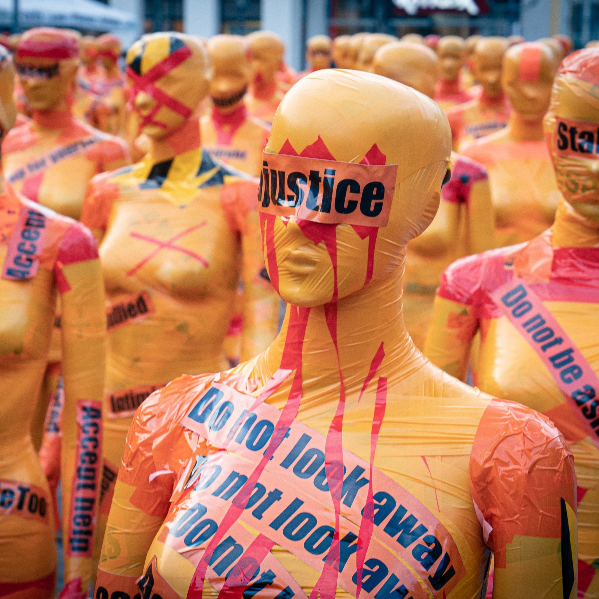 L'image montre un vaste rassemblement de silhouettes humaines couvertes de plastique coloré. Chaque silhouette est peinte en orange vif et ornée de mots et de slogans en noir et en rouge, évoquant des thèmes sociaux et des mouvements de droits civiques comme #MeToo et la justice. Certains mots sont barrés ou soulignés pour accentuer leur impact. L'atmosphère dégage un sentiment fort d'engagement et de protestation, avec ces figures anonymes qui semblent former un mur de revendication. L'ensemble crée une installation artistique saisissante, incitant à la réflexion sur des enjeux sociaux importants.