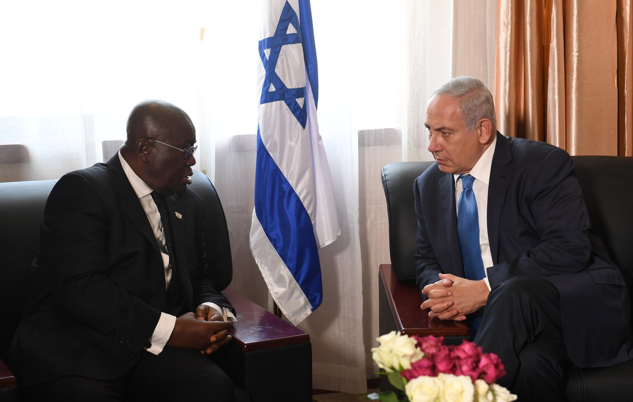 Dans cette image, deux hommes sont assis sur des canapés dans une pièce bien éclairée, entourés de rideaux lumineux. L'un des hommes, à gauche, porte un costume noir et des lunettes, tandis que l'autre, à droite, porte un costume clair et une cravate bleue. Entre eux, on peut voir une petite table avec un bouquet de fleurs aux teintes variées, principalement des roses blanches et des roses. En arrière-plan, se dresse un drapeau israélien, indiquant possiblement un contexte diplomatique. Les deux hommes semblent engagés dans une conversation sérieuse, chacun regardant l'autre avec attention.