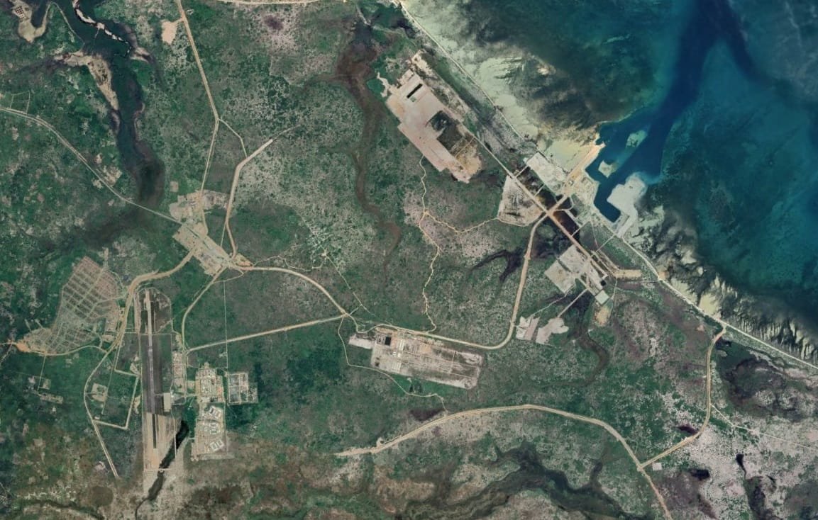 L'image représente une vue aérienne d'une région dont le paysage est marqué par des routes, des structures et des zones déforestées. On peut observer des chemins sinueux qui relient différents points, ainsi que des zones construites qui semblent être des installations industrielles ou militaires. La présence de la mer à droite de l'image ajoute un contraste avec la verdure terrestre, et on peut également voir des étendues d'eau et des zones côtières. Les nuances de couleur vont du vert des zones boisées aux teintes plus claires des infrastructures.